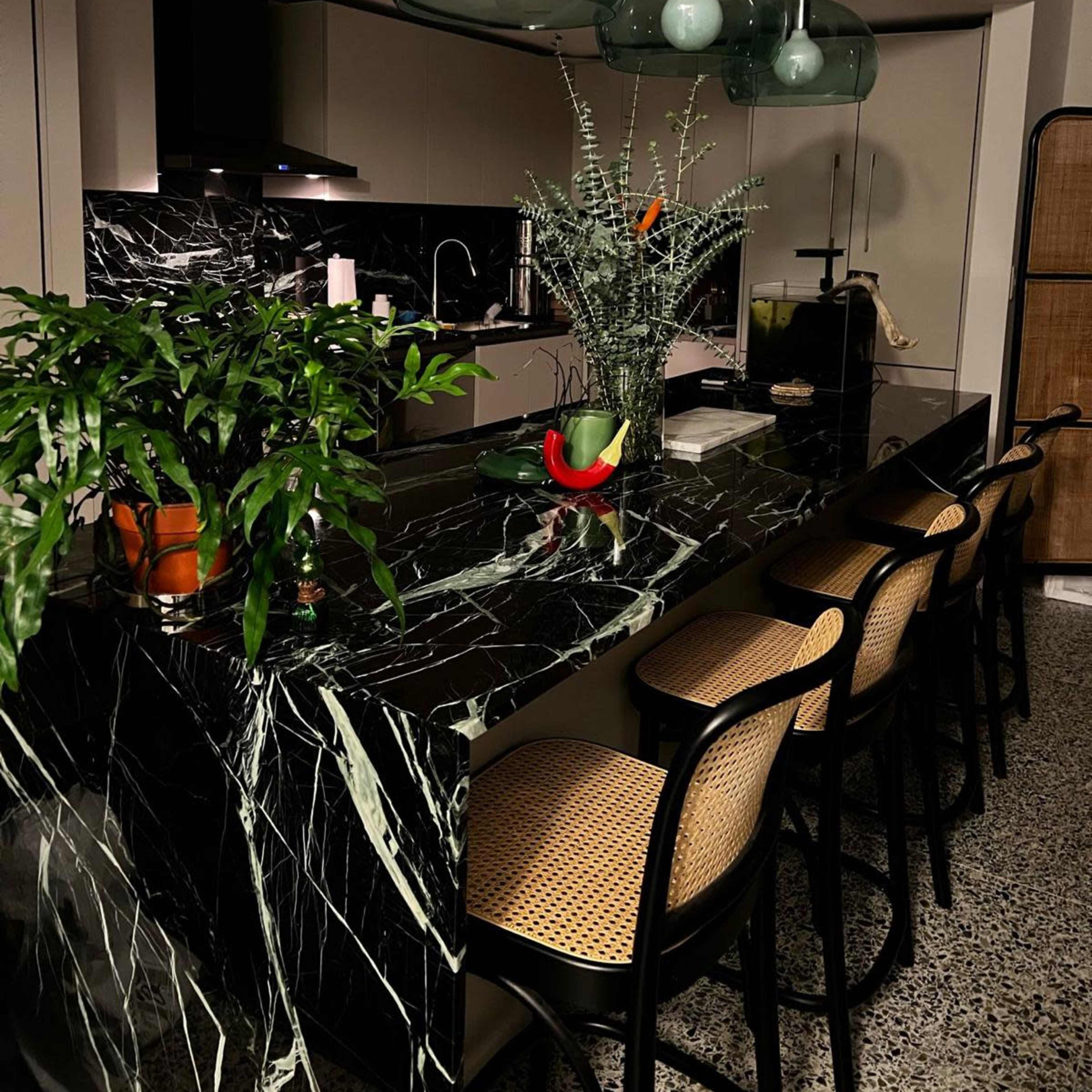 A modern kitchen featuring a black marble countertop, three green pendant lights, and bar stools along the counter, with plants and a fruit bowl as decor.