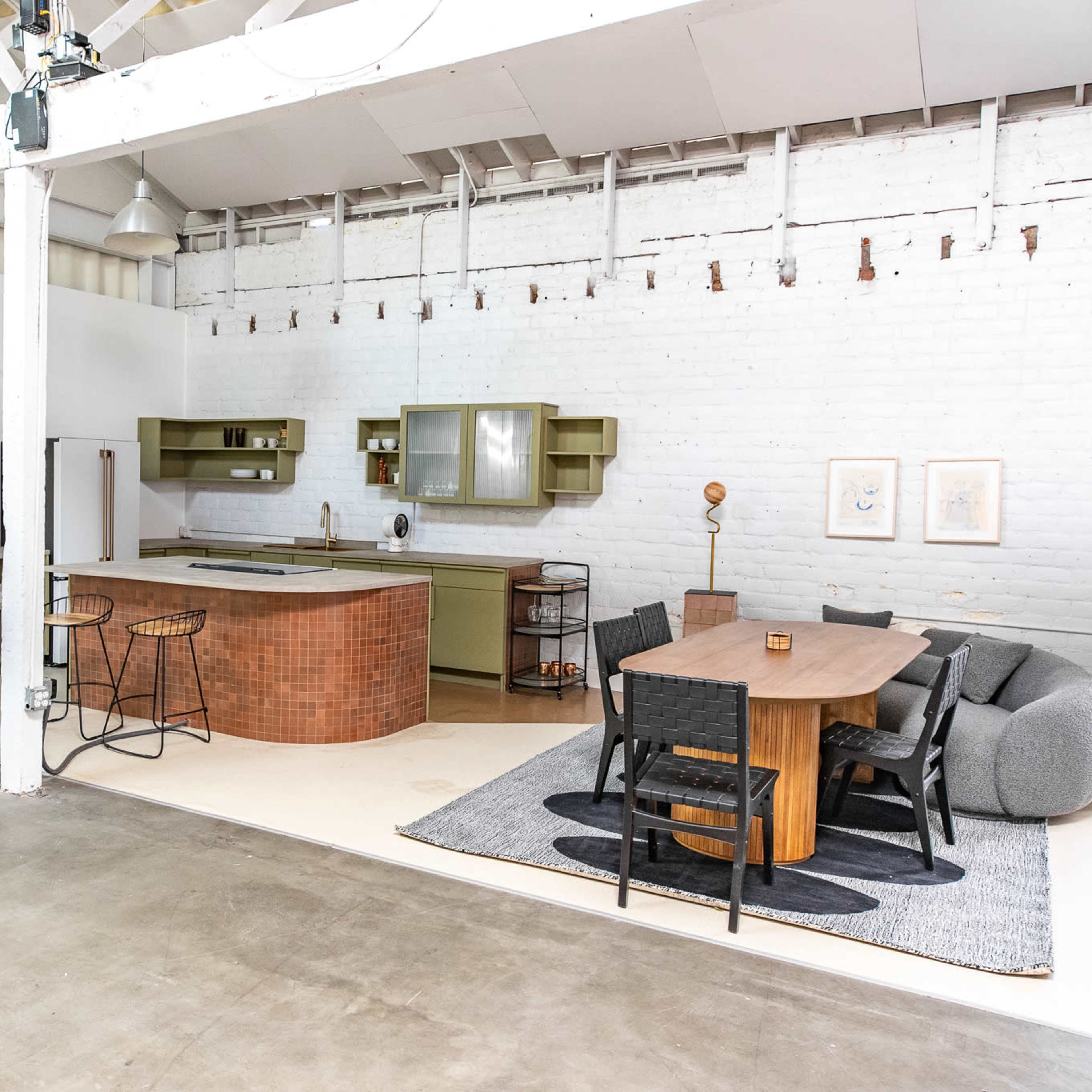 A spacious, modern kitchen and dining area featuring a large wooden table, black chairs, and a kitchen island with green cabinetry against a white brick wall.