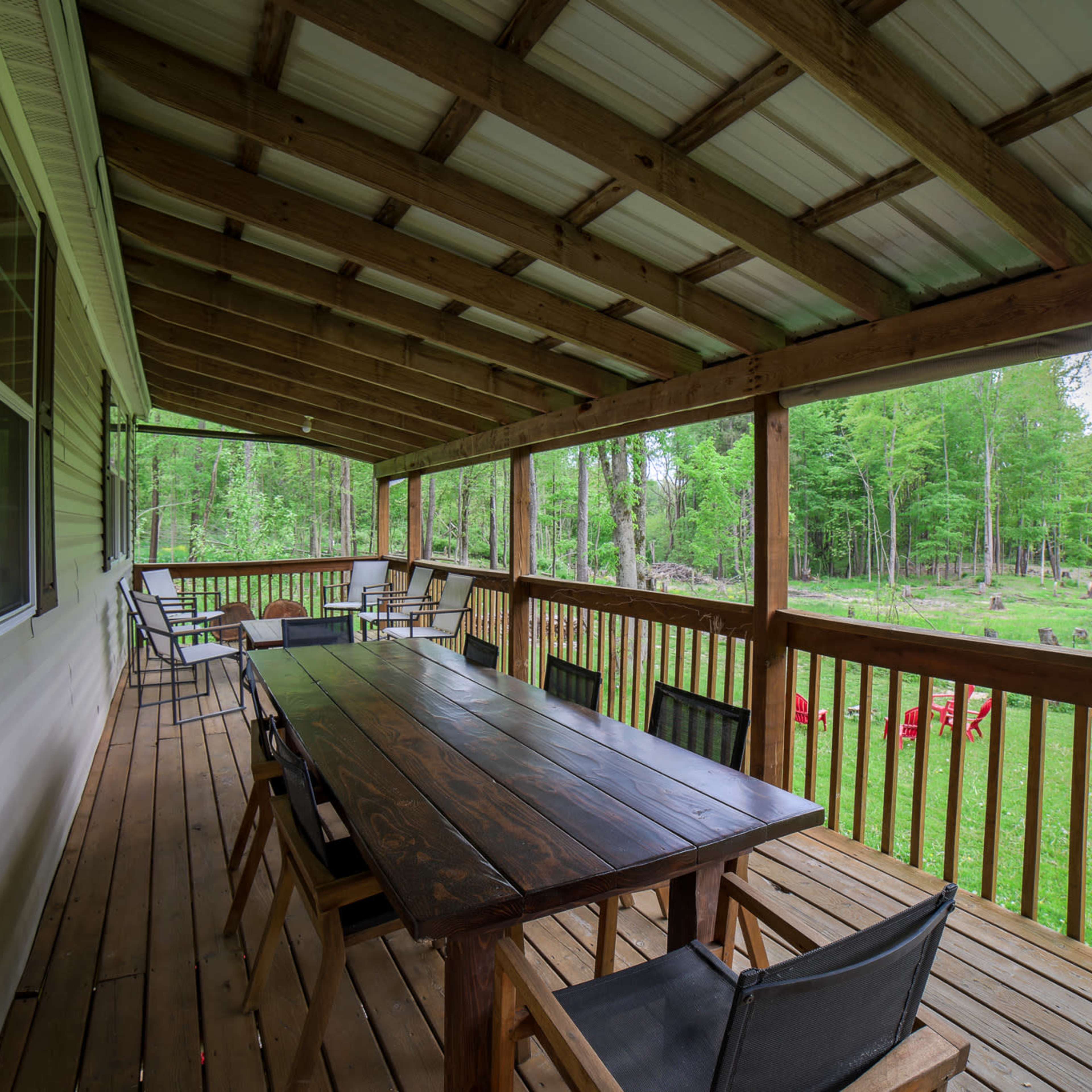 A wooden deck with a long table and chairs under a slanted roof, surrounded by trees and a grassy area.