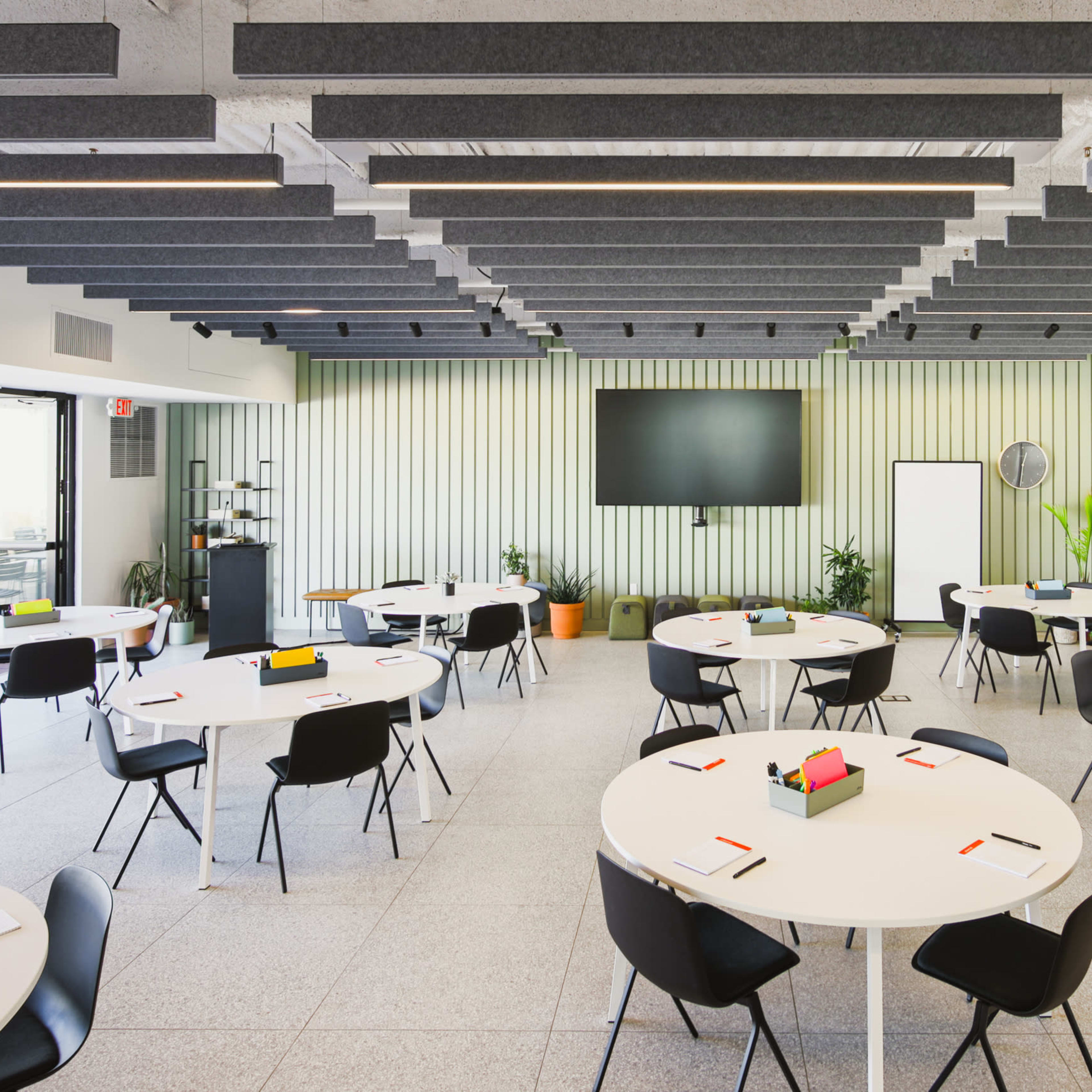 A modern conference room features multiple round tables set with stationery, surrounded by sleek black chairs and equipped with a large screen at one end.