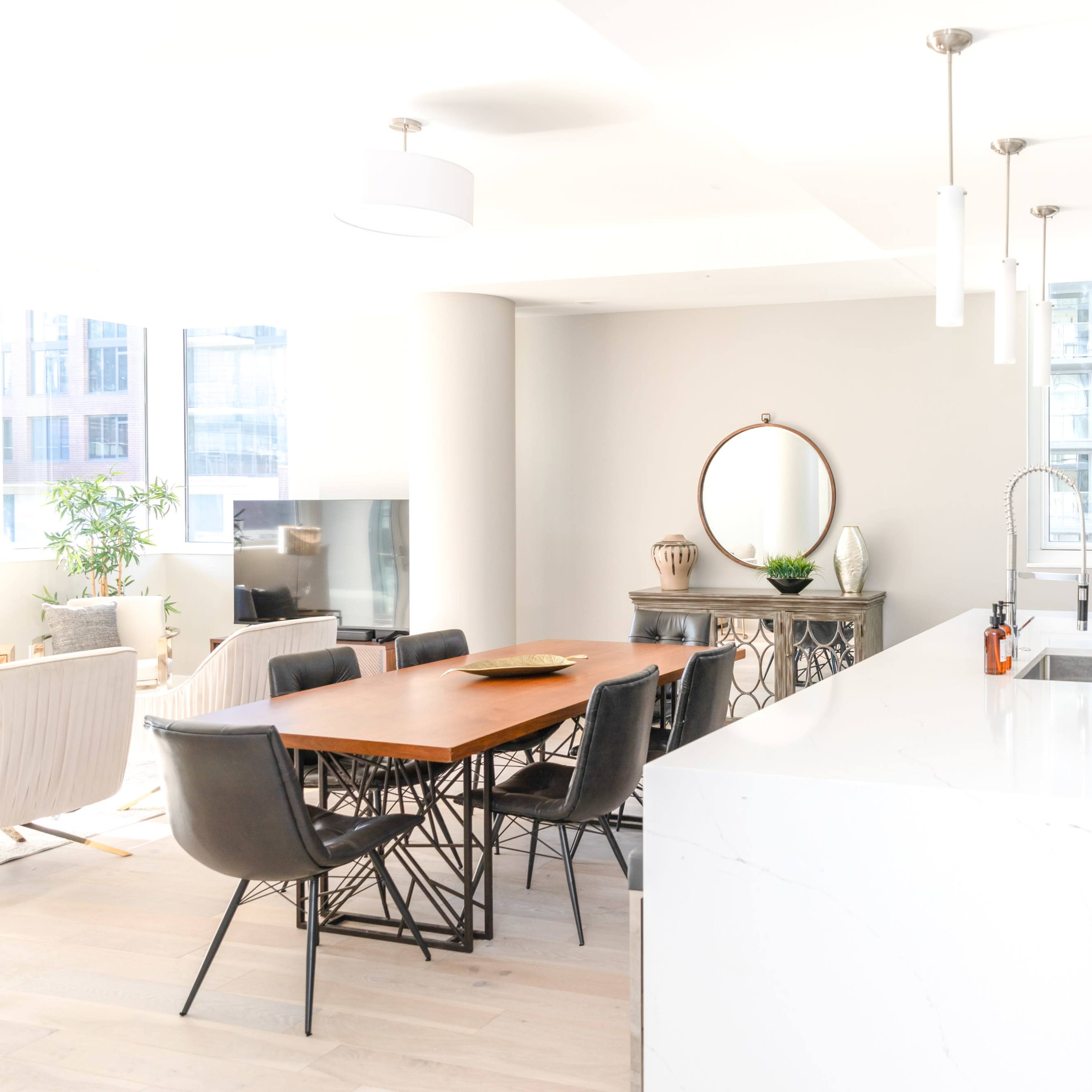 A modern kitchen and dining area feature a white island, a wooden dining table with black chairs, and large windows allowing natural light to fill the space.