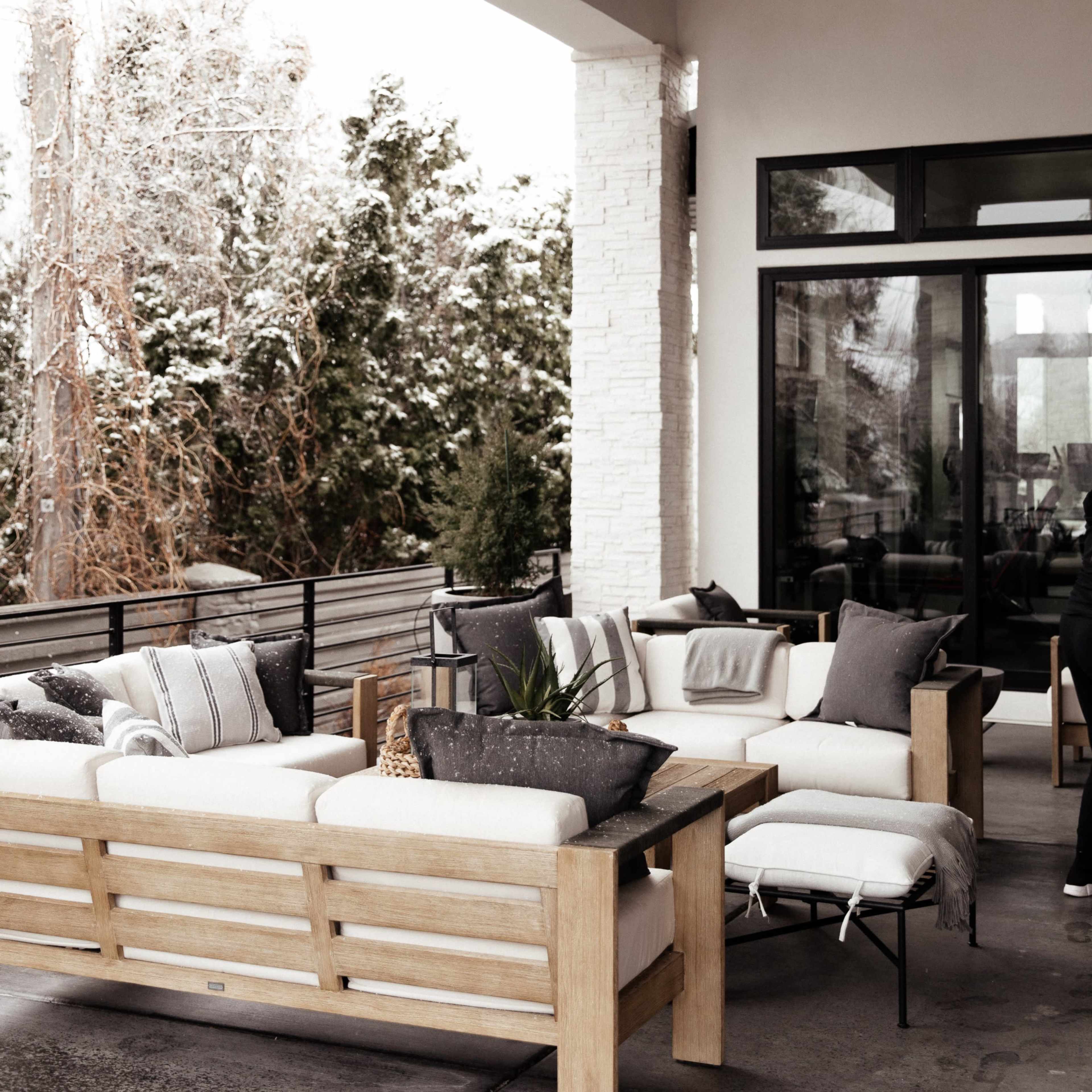 A covered patio features wooden furniture arranged around a low table, with decorative pillows and a backdrop of greenery and a snowy landscape.