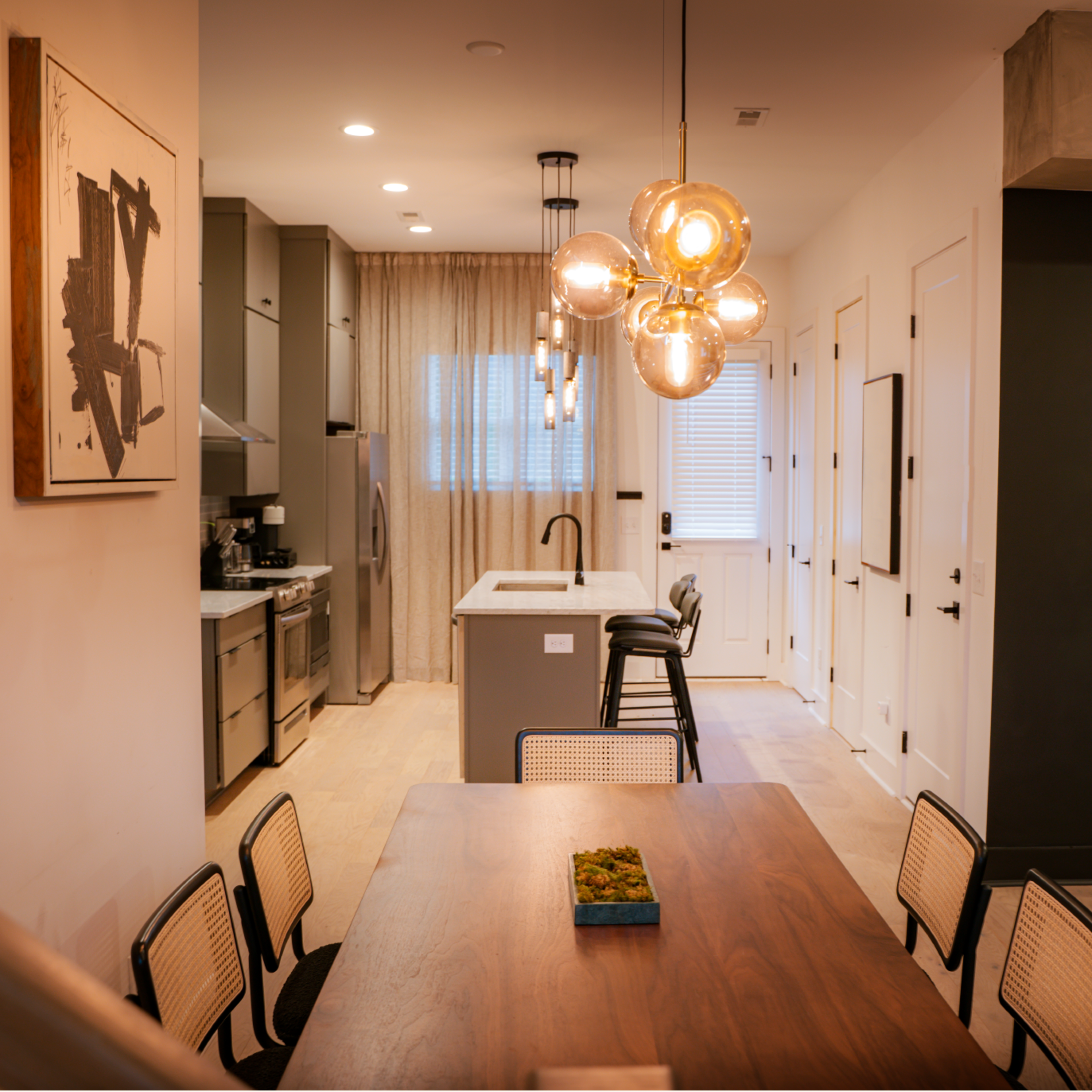 A modern kitchen and dining area with a wooden dining table, stylish lighting fixtures, and a view of the kitchen equipped with stainless steel appliances.