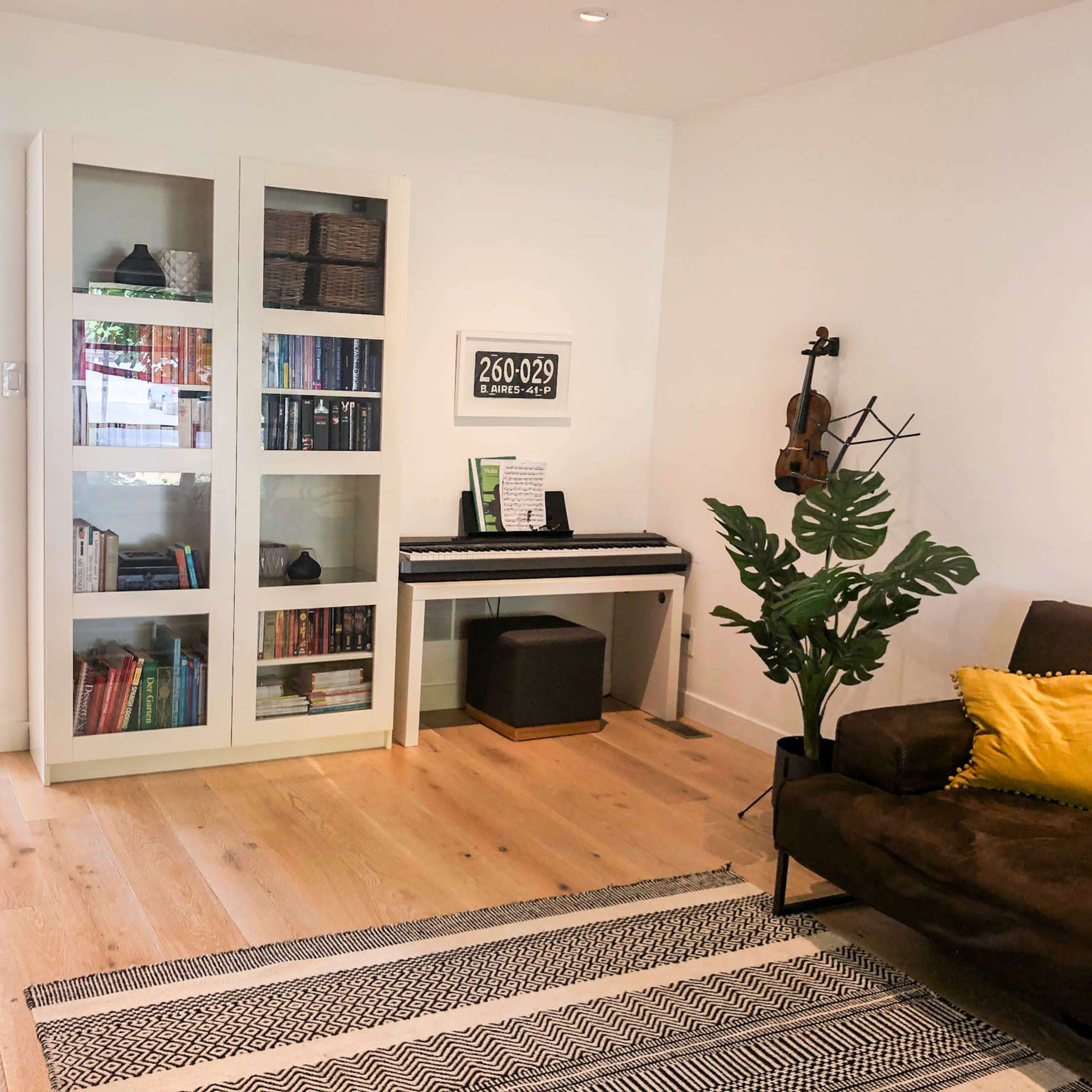 The room features a brown sofa, a black and white striped rug, a bookshelf filled with books, a piano with sheet music, and a potted plant.
