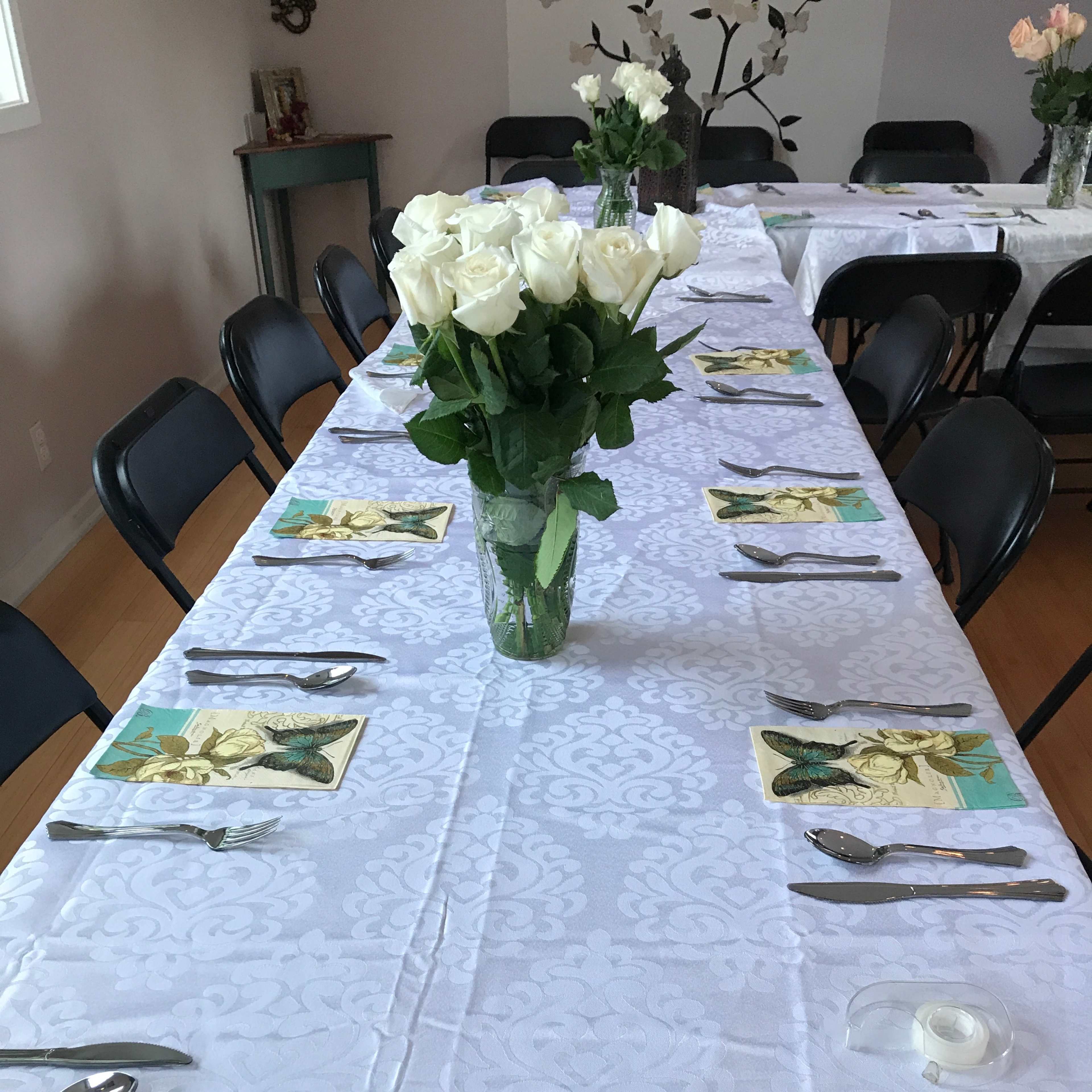 A long table is set with white tablecloths, black chairs, and a vase of white roses at the center, surrounded by plates, utensils, and napkins.