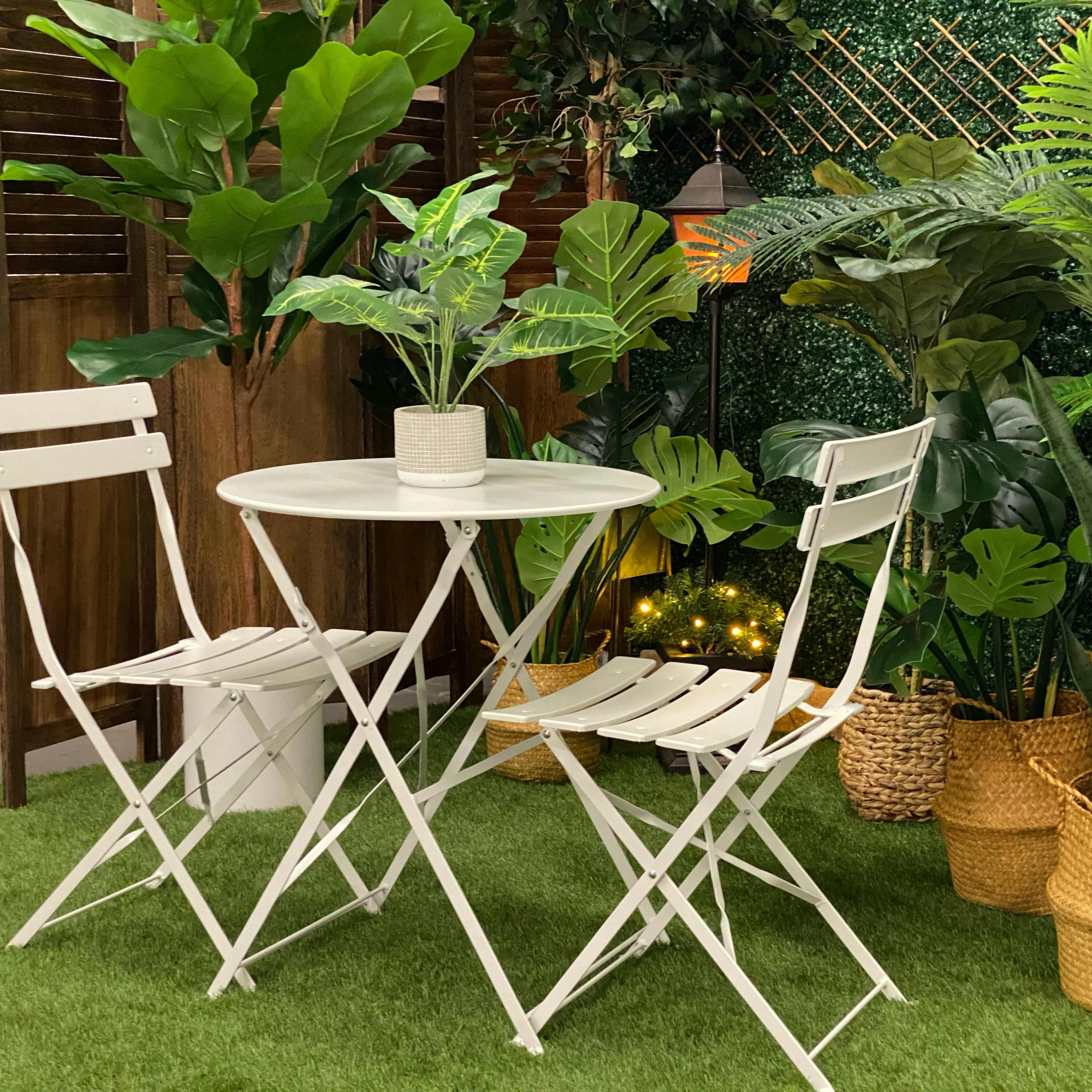 A small white table with two folding chairs is surrounded by various potted plants in a green, artificial grass setting.