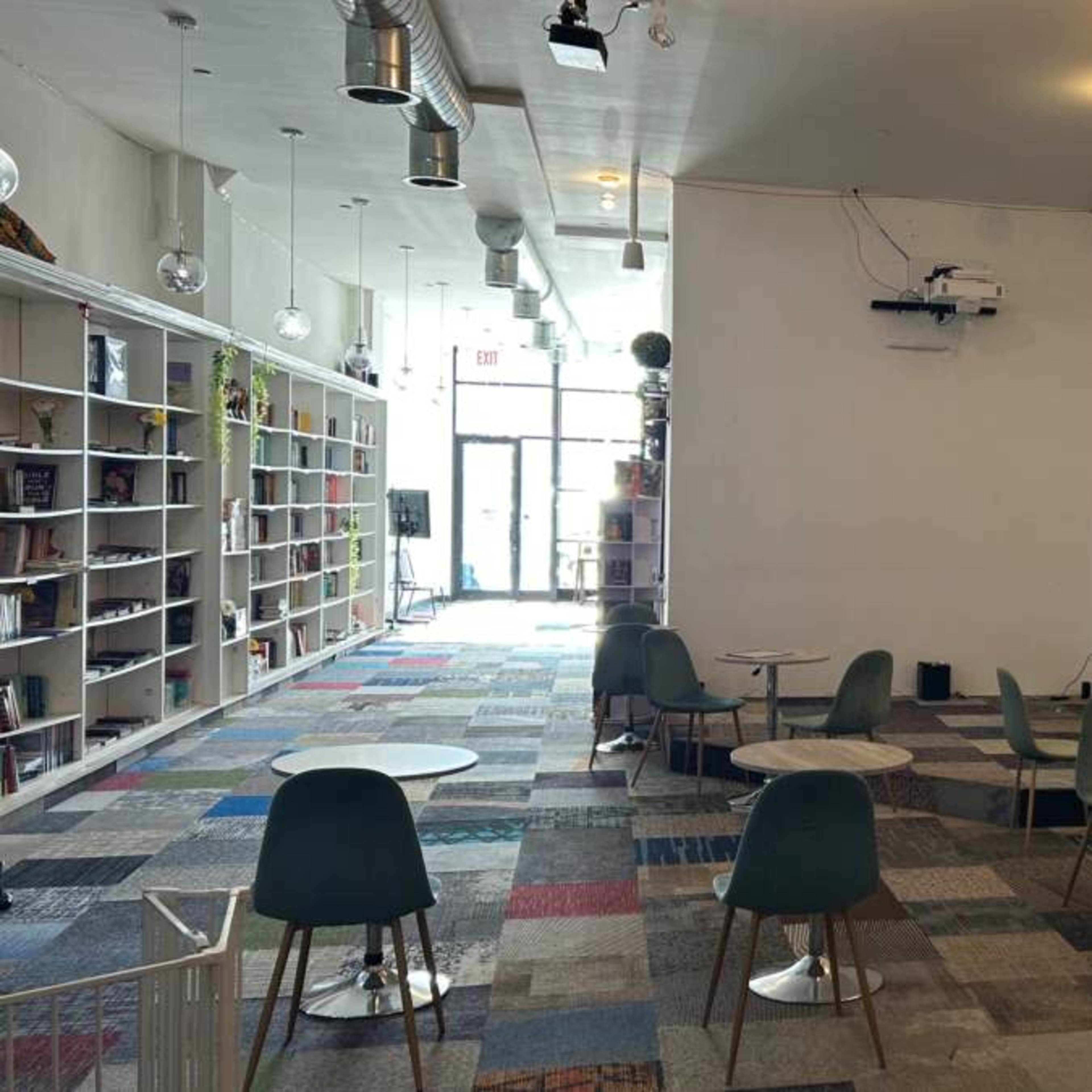 A spacious, minimalist café with several small tables and green chairs, a projector screen on the wall, and a bookshelf along one side.