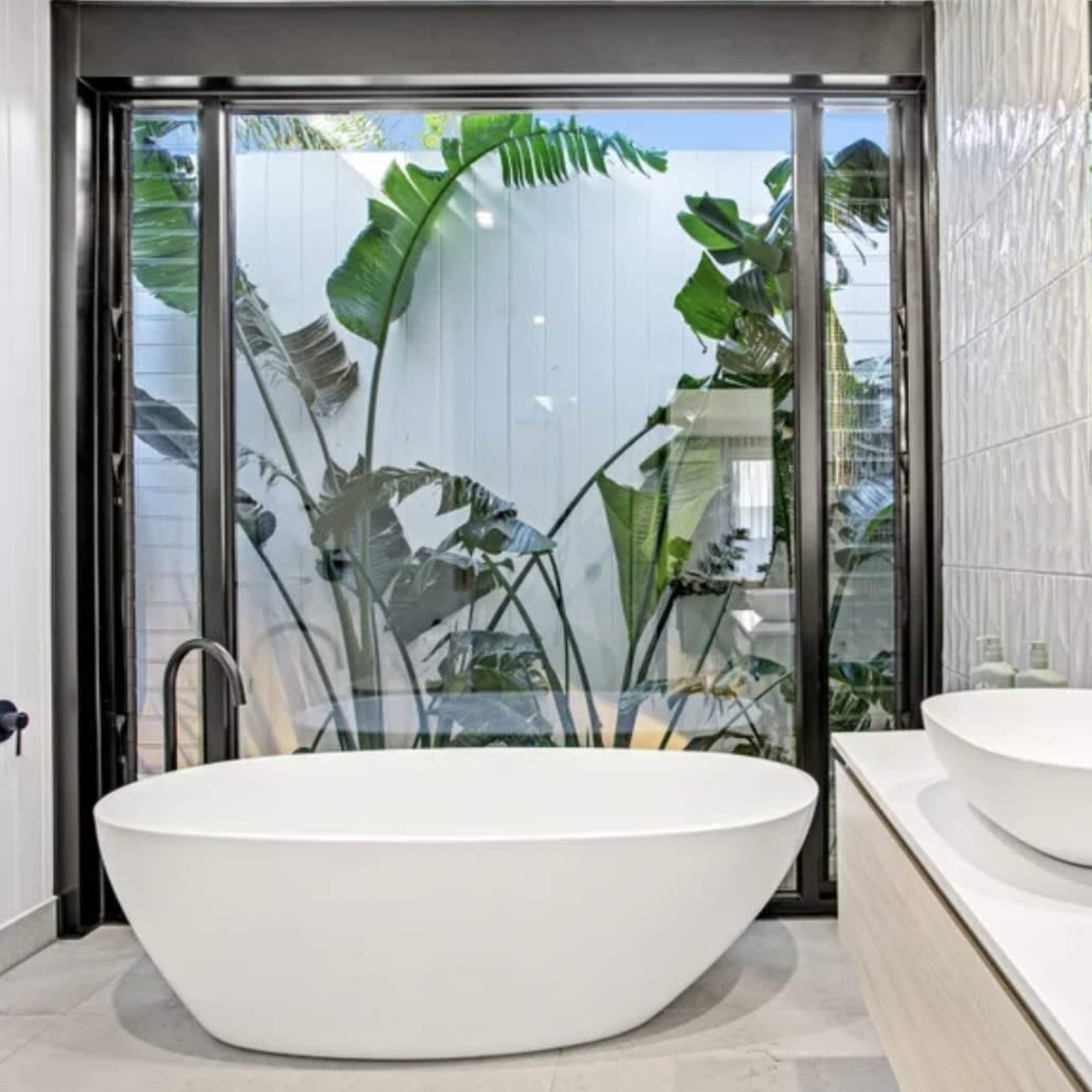 A modern bathroom with a freestanding white bathtub, a double sink vanity, and tall glass windows showcasing greenery outside.