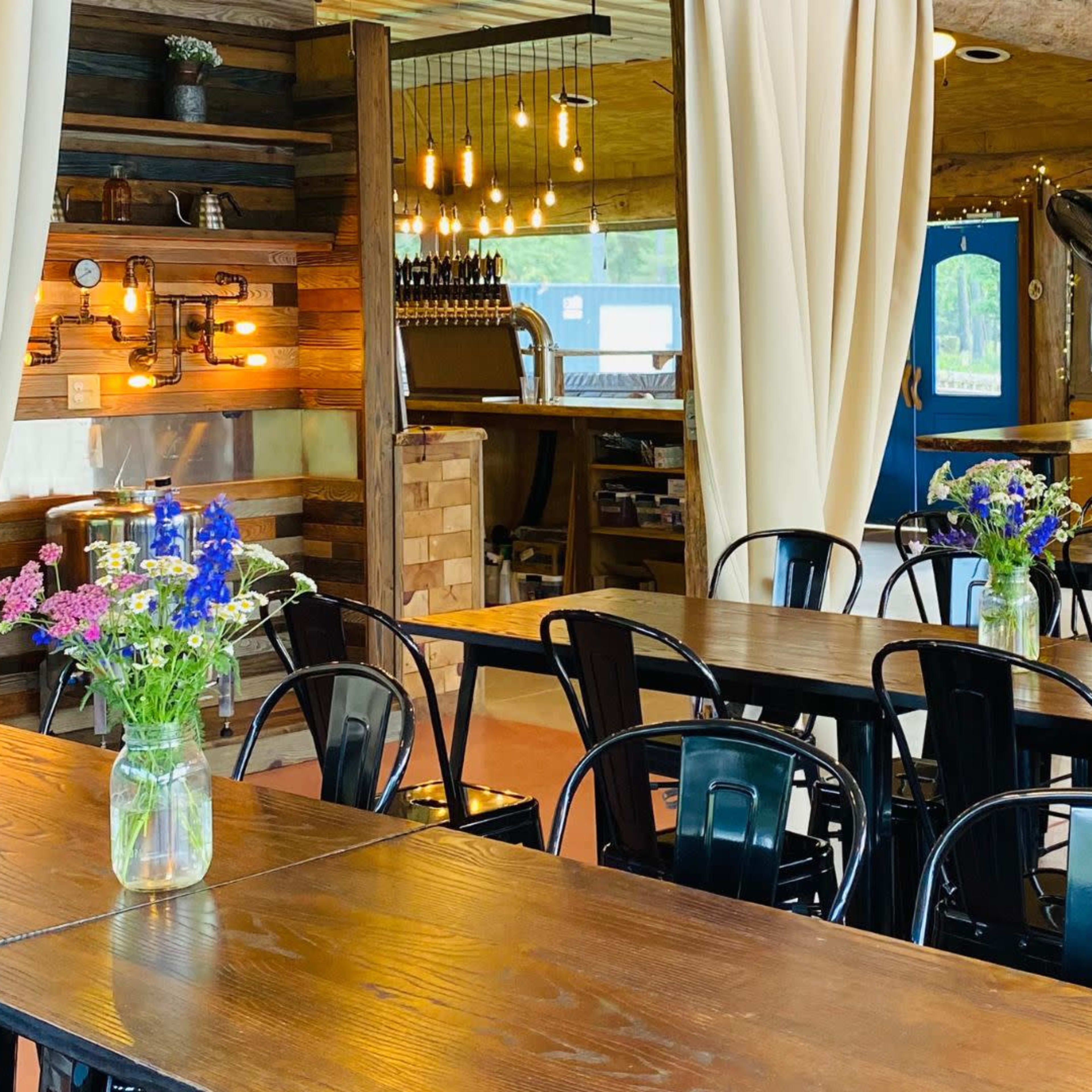 A dining area with wooden tables, black metal chairs, and flower vases on each table in a well-lit setting.