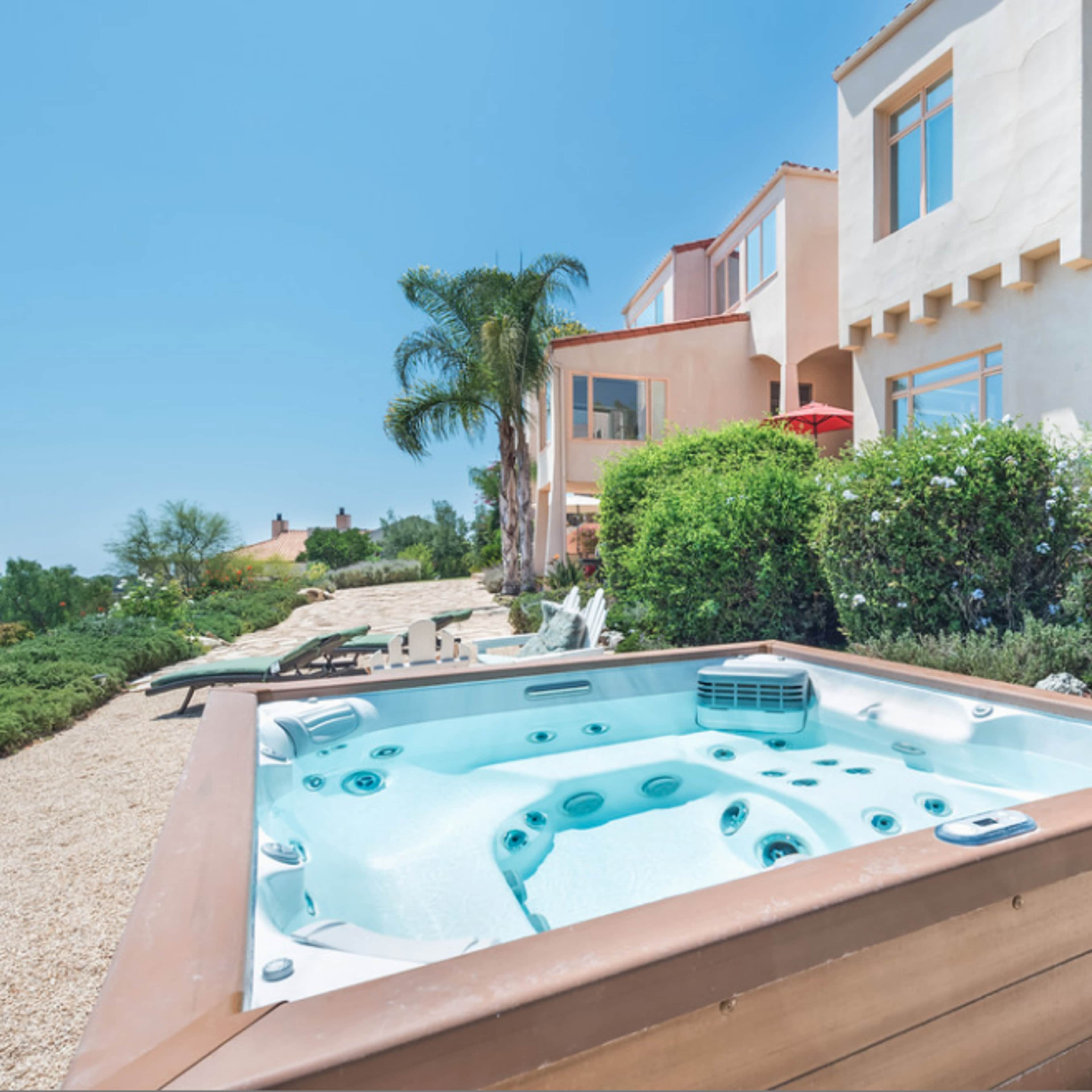 A hot tub sits in a landscaped yard with a view of a house and surrounding greenery.