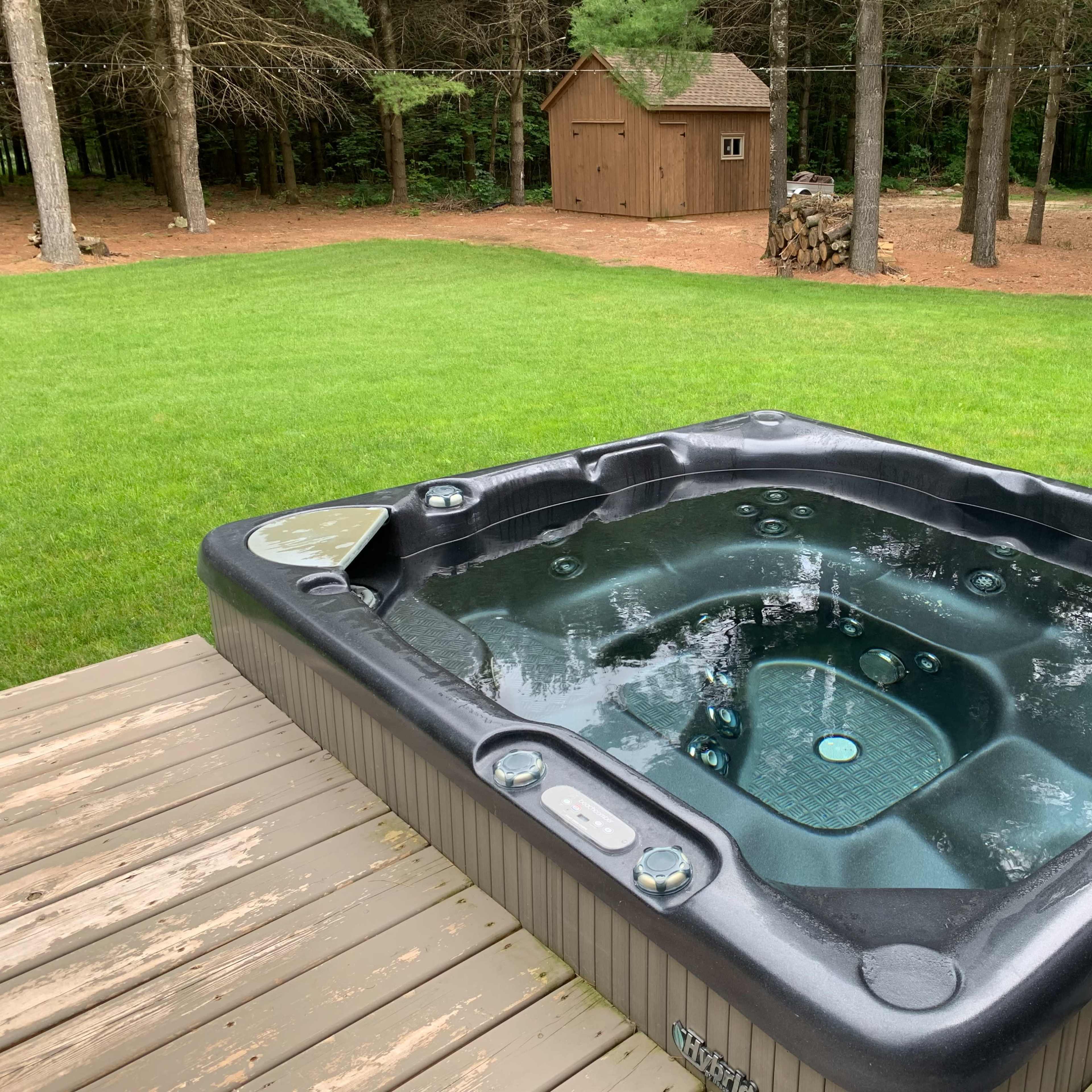 A hot tub sits on a wooden deck in a grassy yard, with a shed and trees in the background.