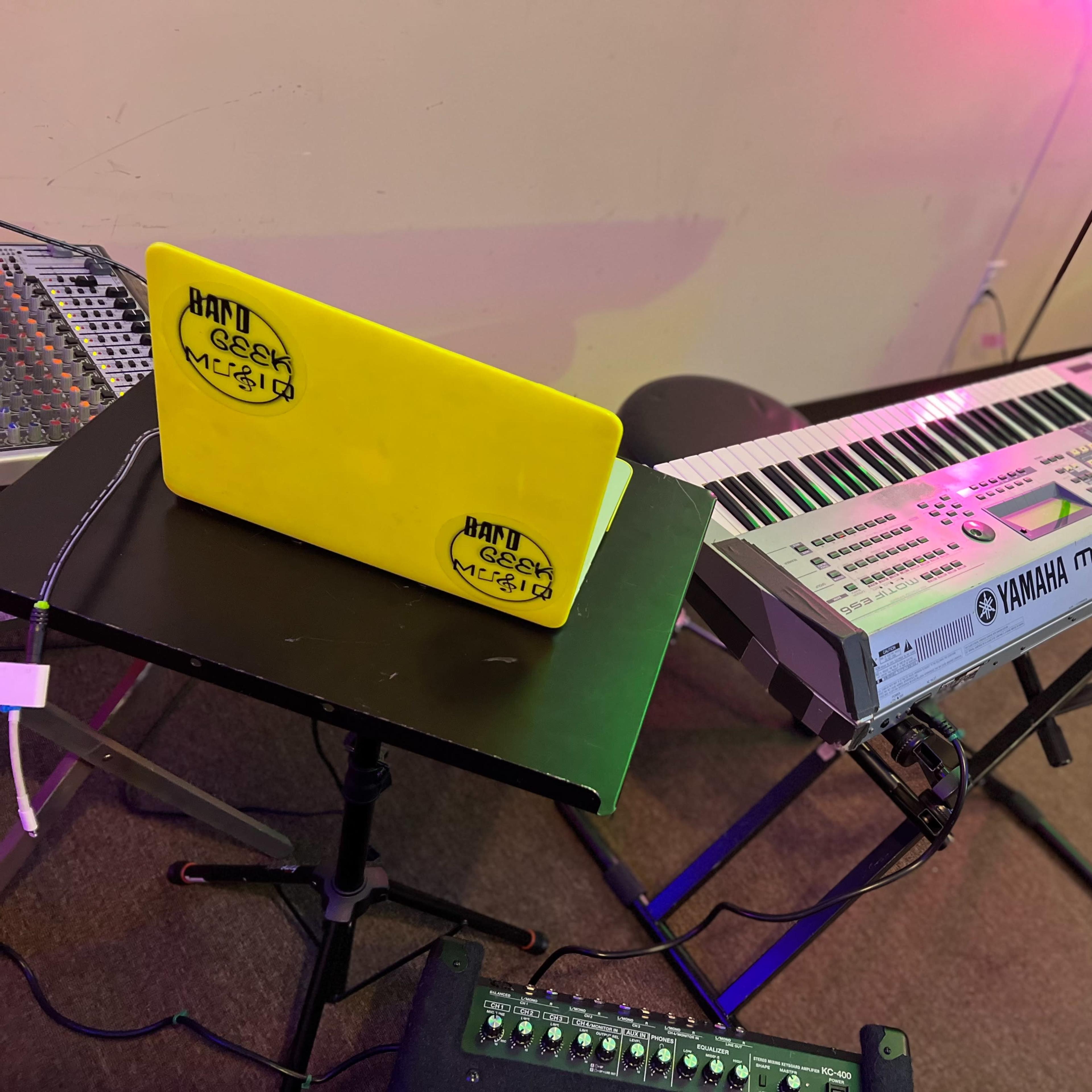 A yellow laptop with stickers is placed on a black music stand next to a Yamaha Motif ES keyboard, both set up in a room with colorful lighting.