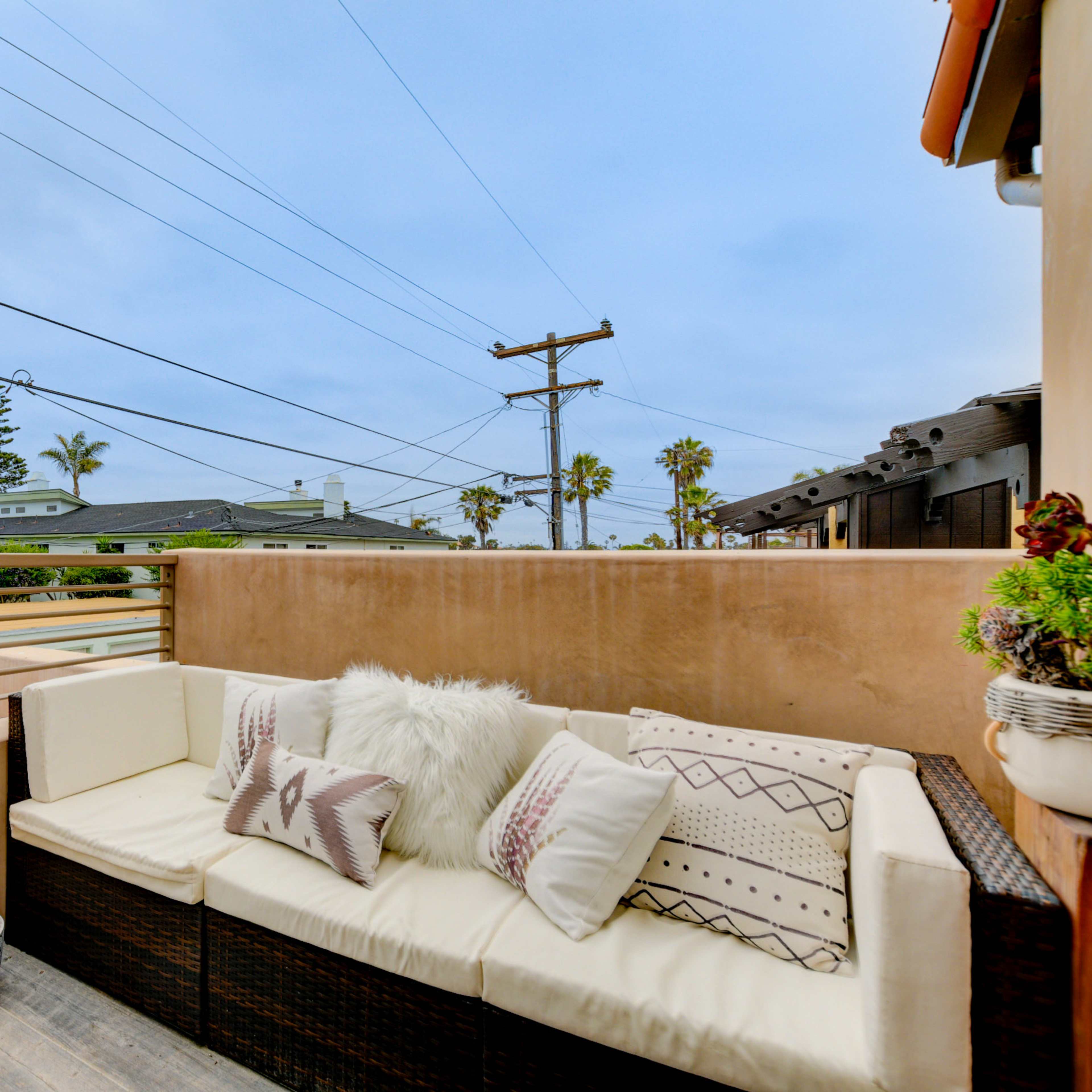 A cozy outdoor seating area is arranged with cushions, surrounded by palm trees and power lines against a cloudy sky.