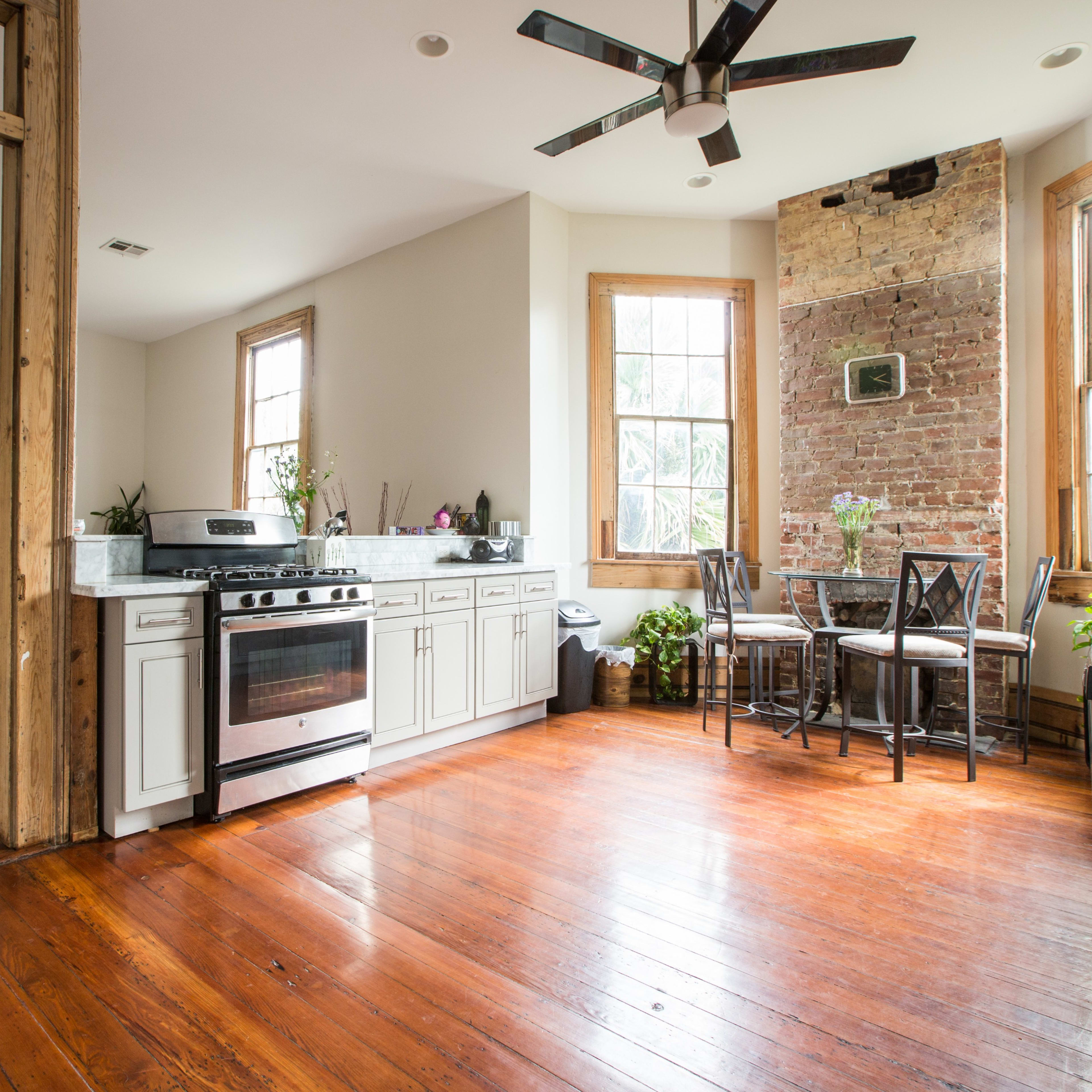 A kitchen with a stove, cabinets, and a dining area featuring a table and chairs, adjacent to a living space with wooden floors and large windows.