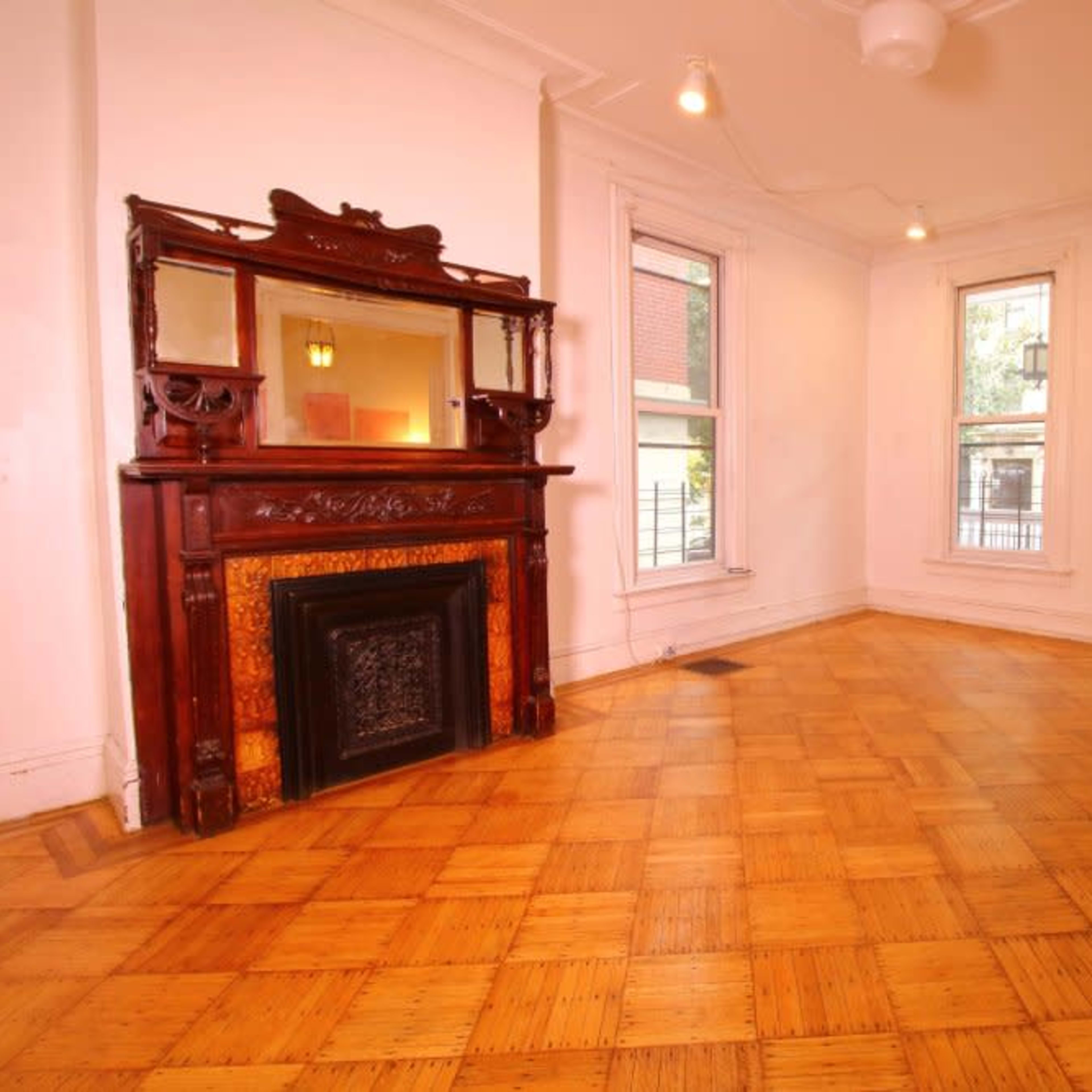 A brightly lit room with polished parquet flooring, featuring a wooden fireplace mantel and a mirror above it.