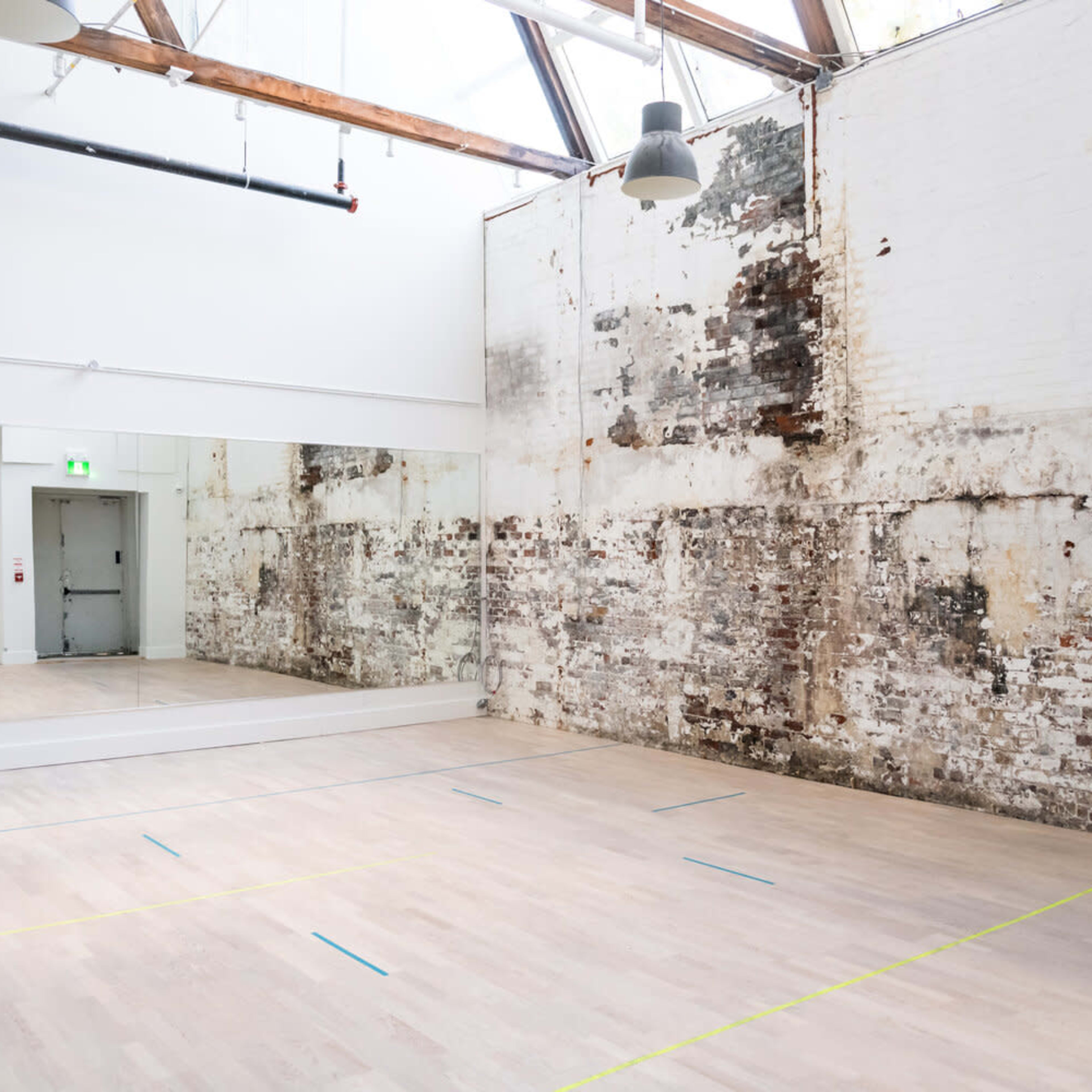 A spacious dance studio with wooden floors, a large mirrored wall, and unfinished brick walls, illuminated by overhead lighting and natural light from a skylight.