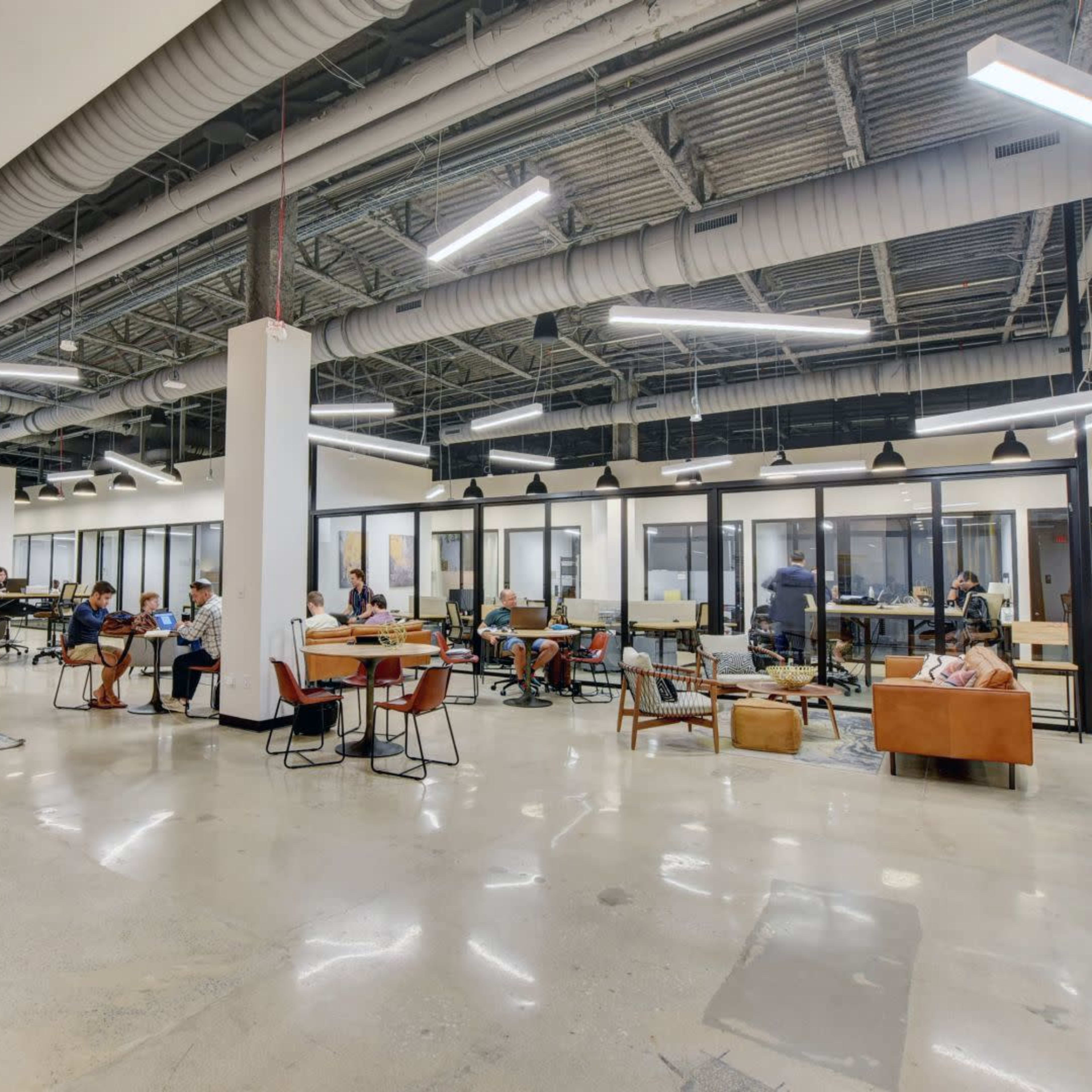 A modern office space with several workstations, lounge areas, and glass-enclosed meeting rooms, populated by individuals engaged in various activities.