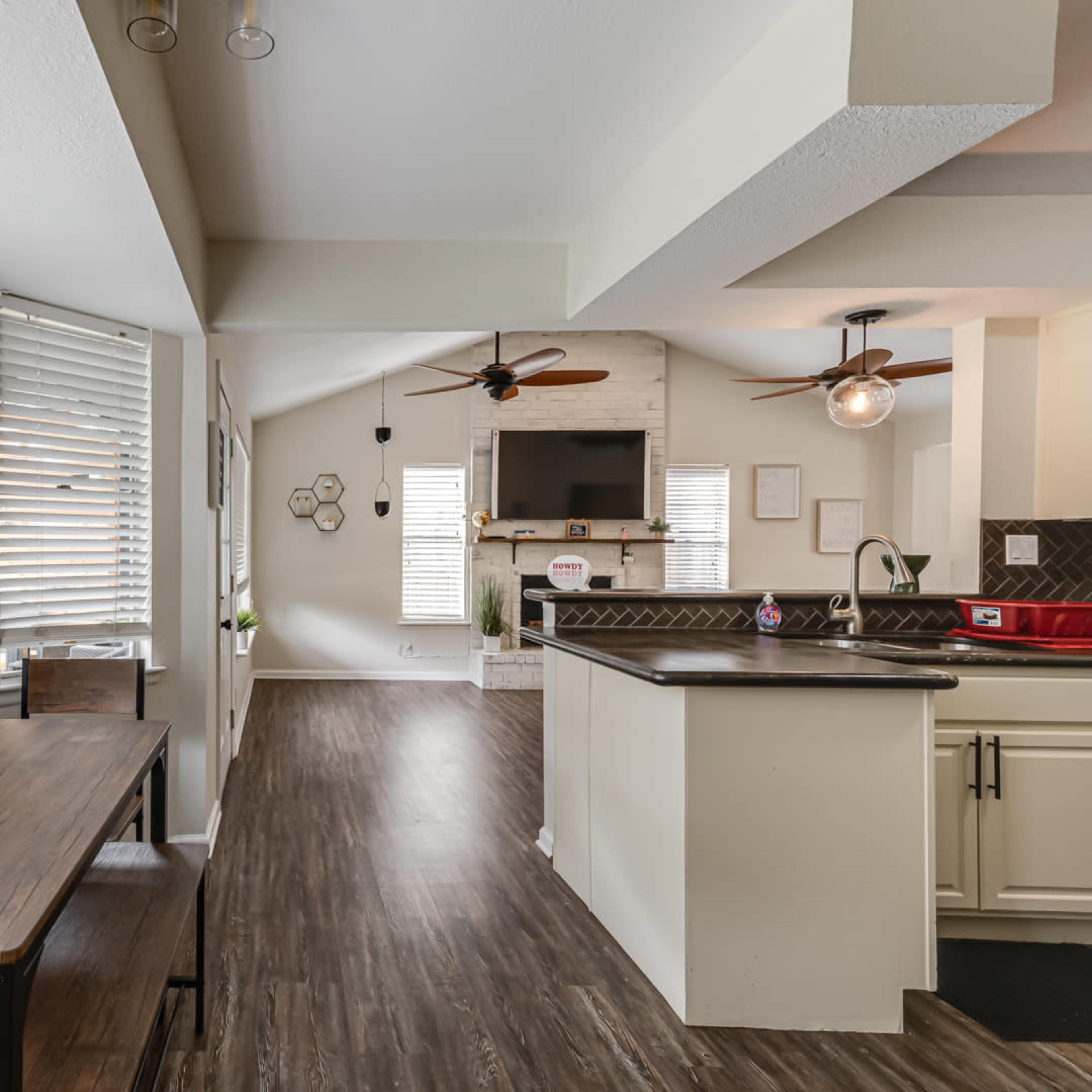 A modern kitchen and dining area feature a wooden table near large windows, with a living space that includes a television and a cozy seating arrangement.