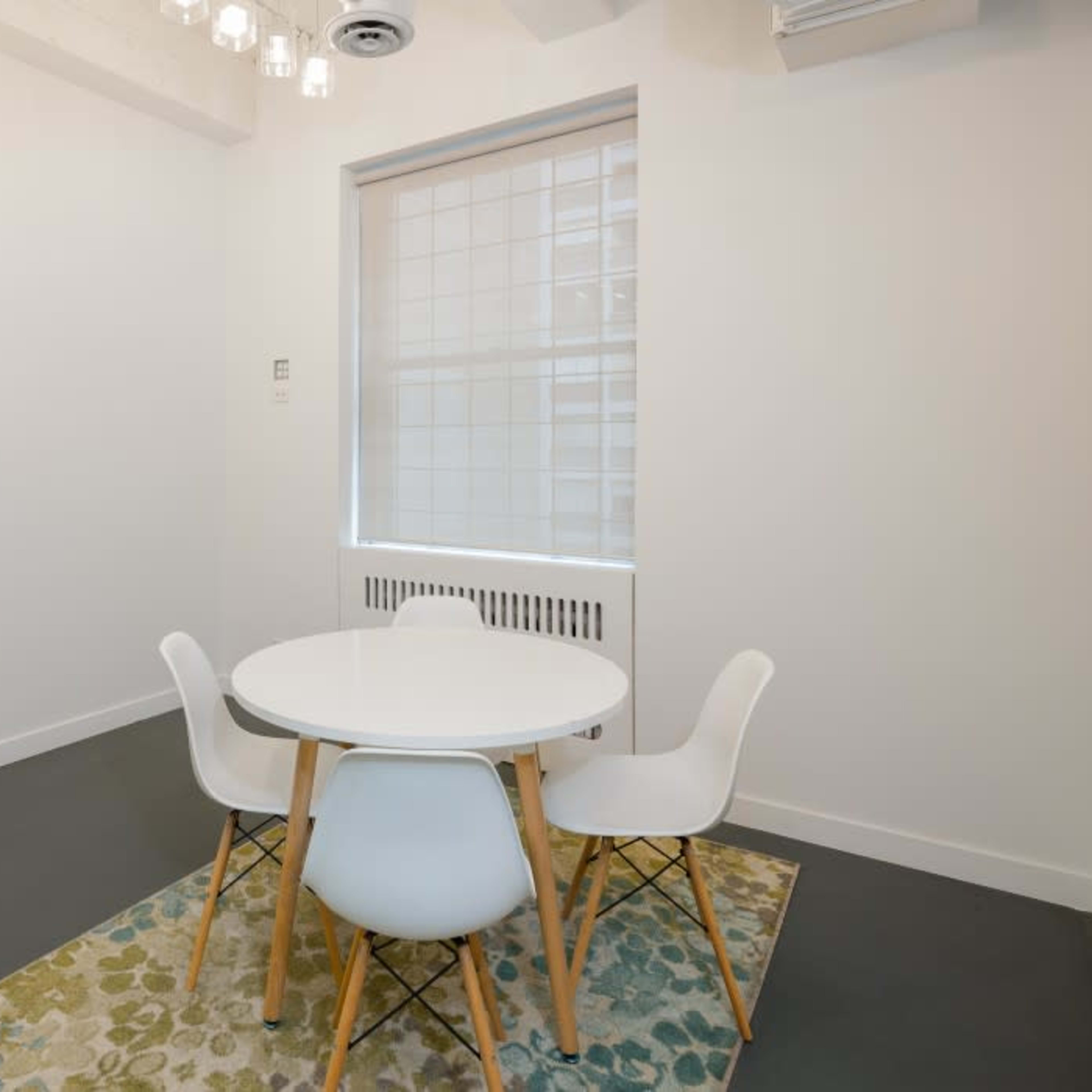 A minimalistic dining area with a round white table and four white chairs on a patterned rug in a bright, light-colored room.