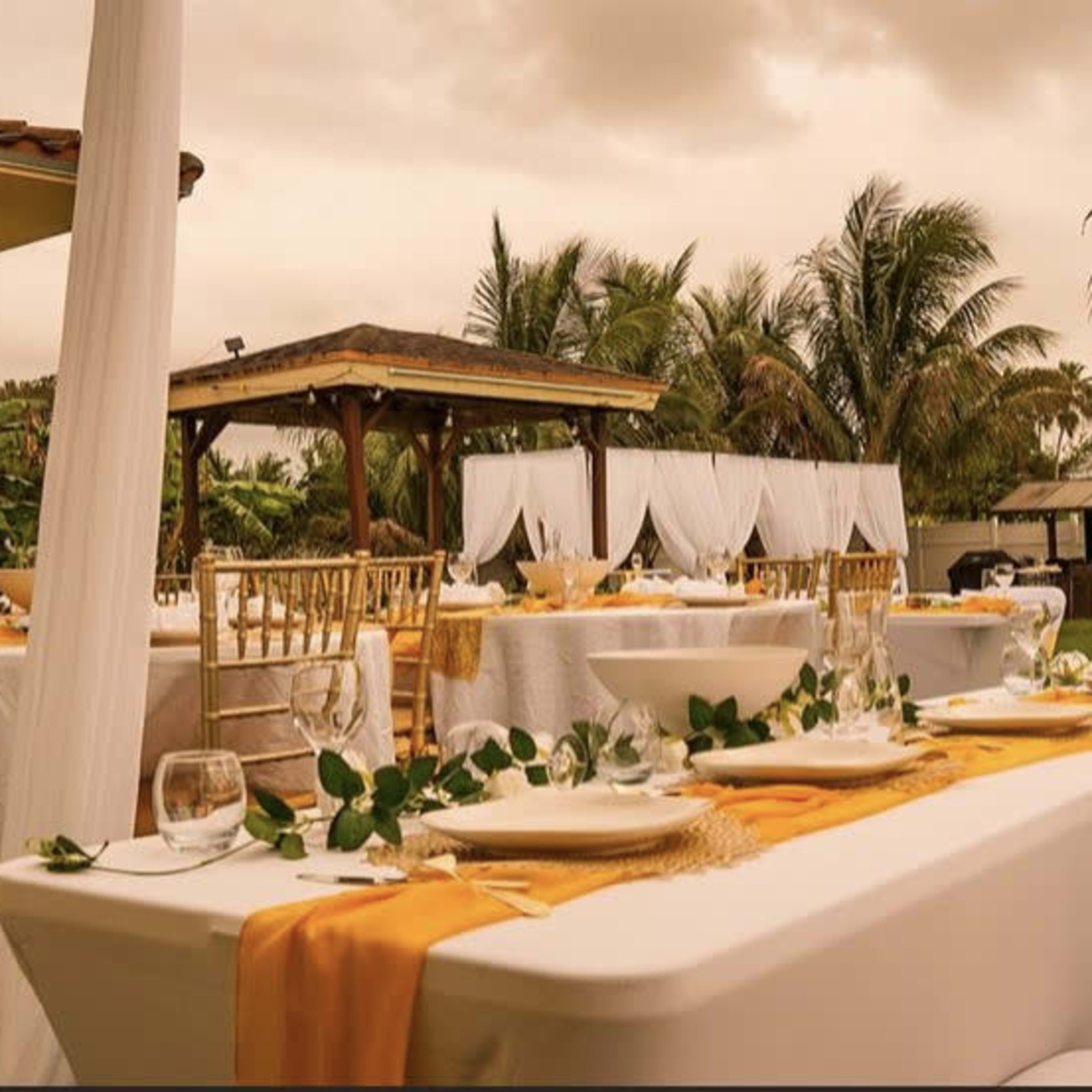 A spacious outdoor dining setup features elegantly arranged tables with white tablecloths, golden accents, and greenery, surrounded by palm trees and a gazebo.