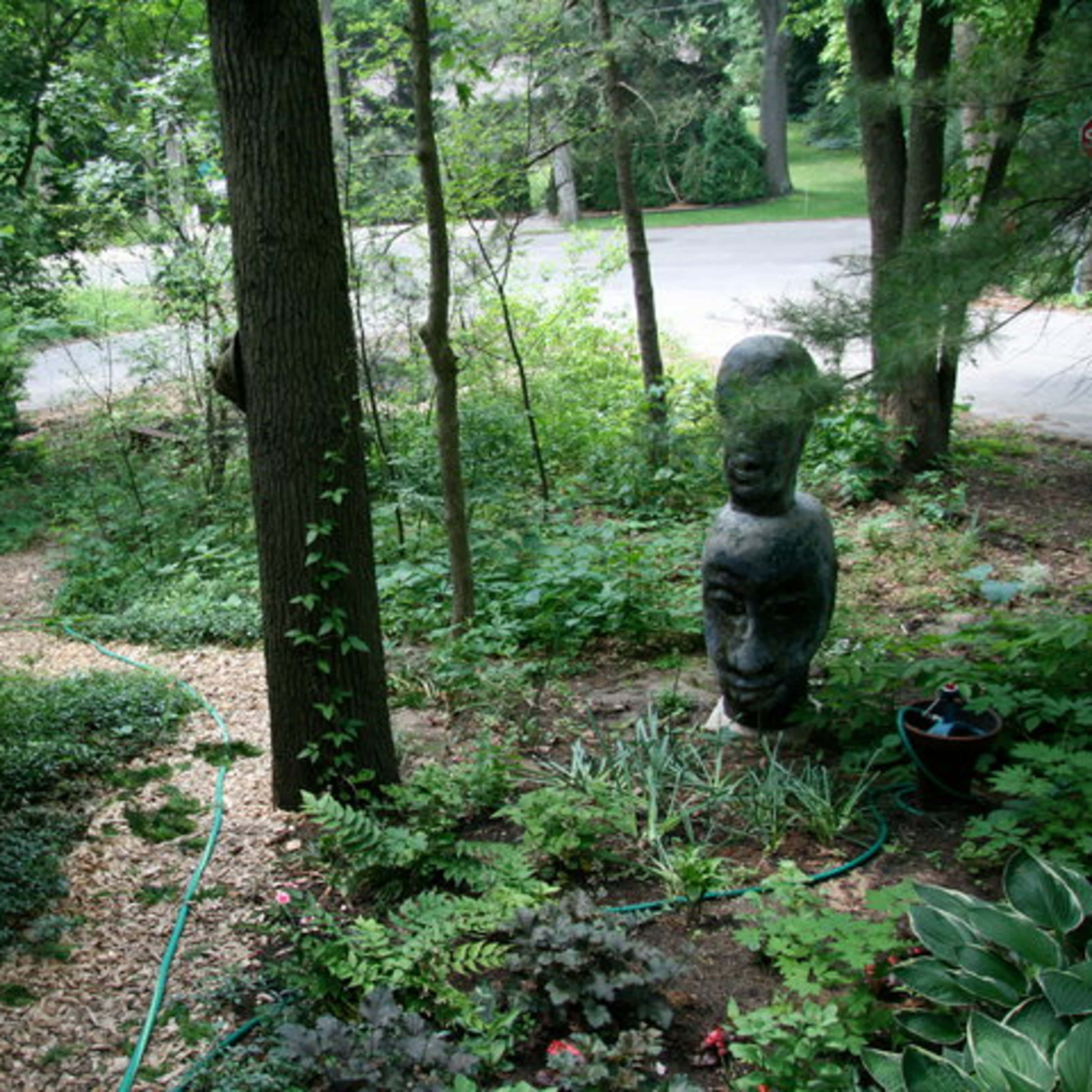 A lush, green garden with a pathway leading through dense foliage and a large wooden sculpture positioned among the trees.