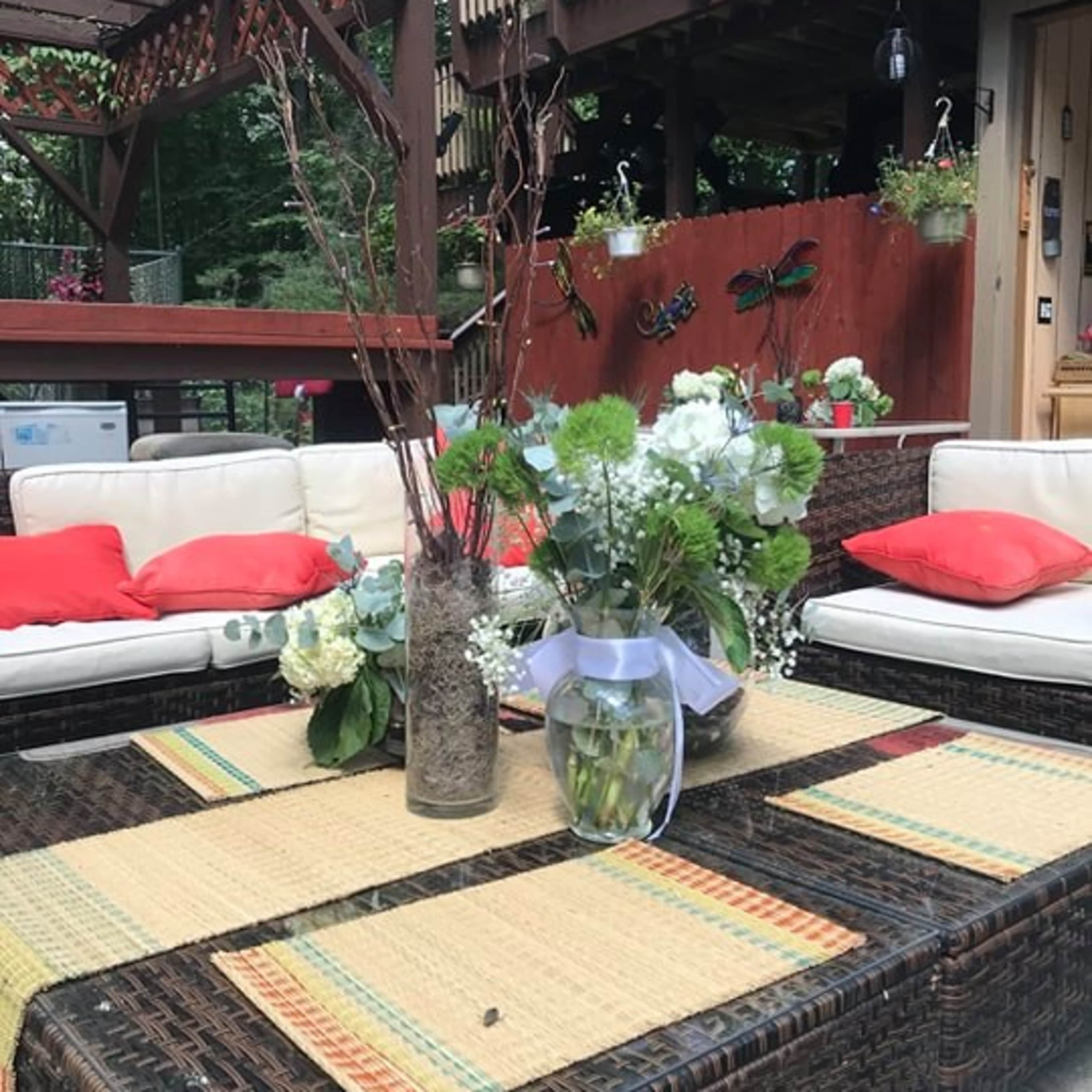 A patio setting features a table adorned with floral arrangements, surrounded by cushioned seating and a wooden deck in the background.