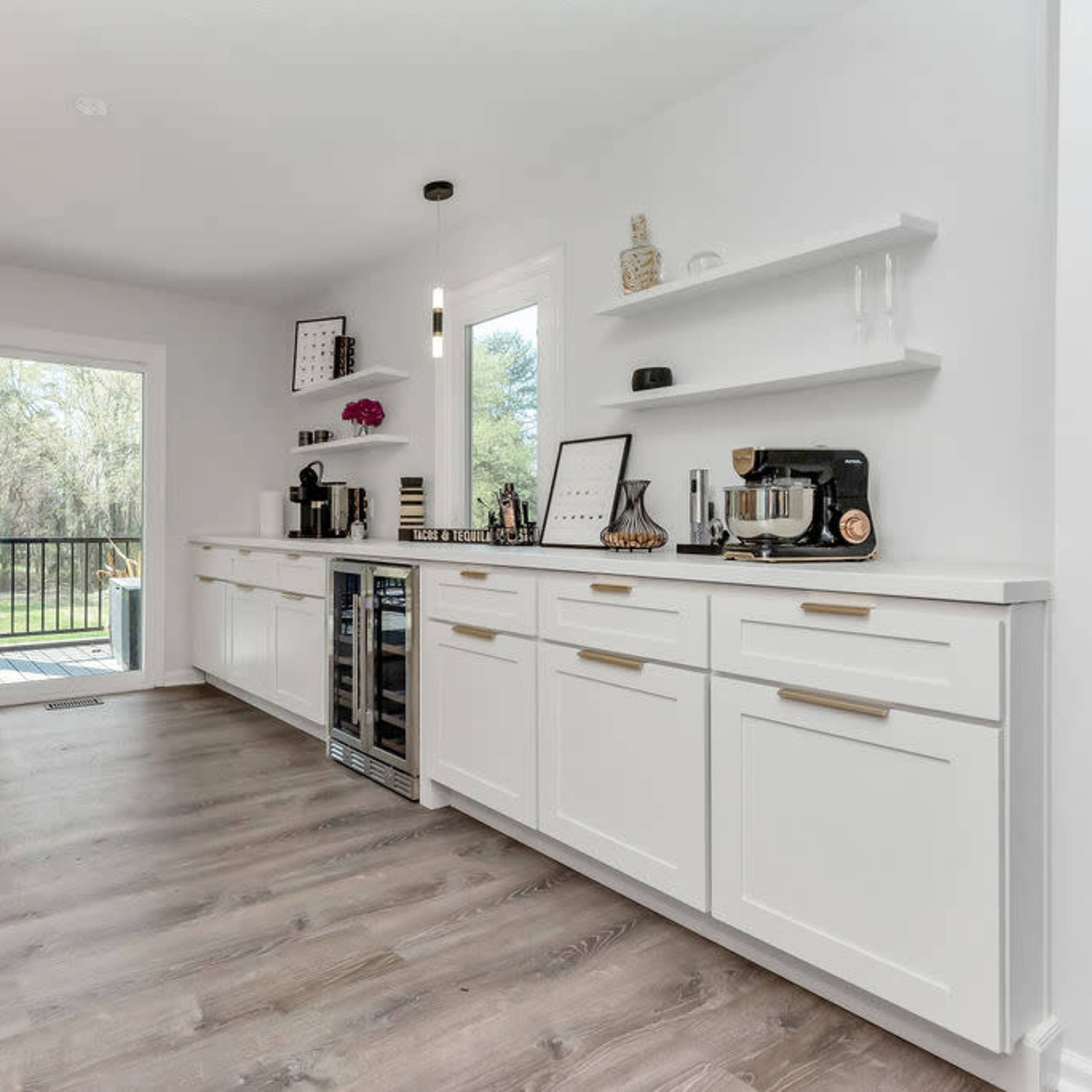 A modern kitchen with white cabinetry, a countertop, and stainless steel appliances, featuring a small dining area with gold bar stools and large windows overlooking a deck.