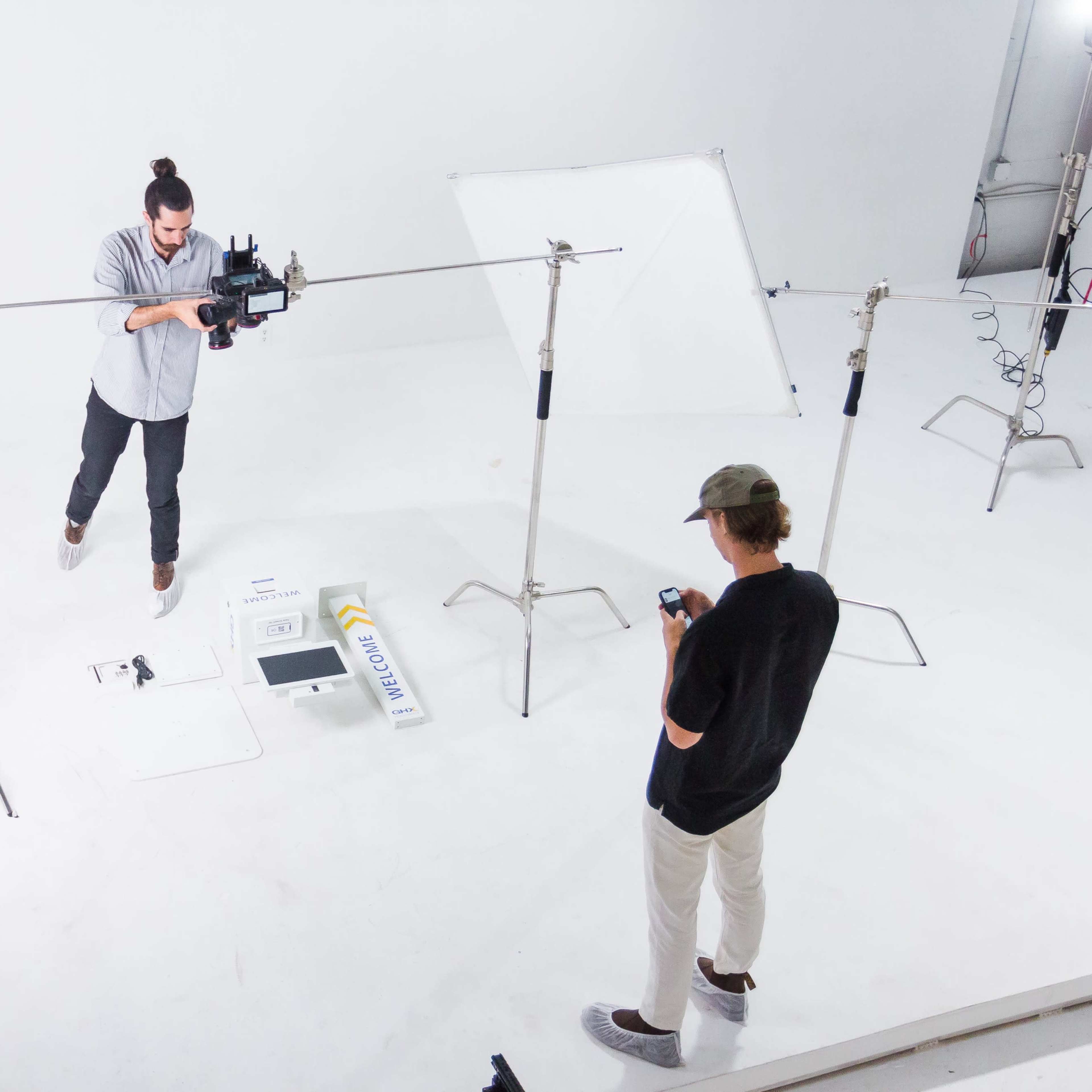 Two individuals are in a photography studio setting, one adjusting a camera on a tripod while the other checks their phone.