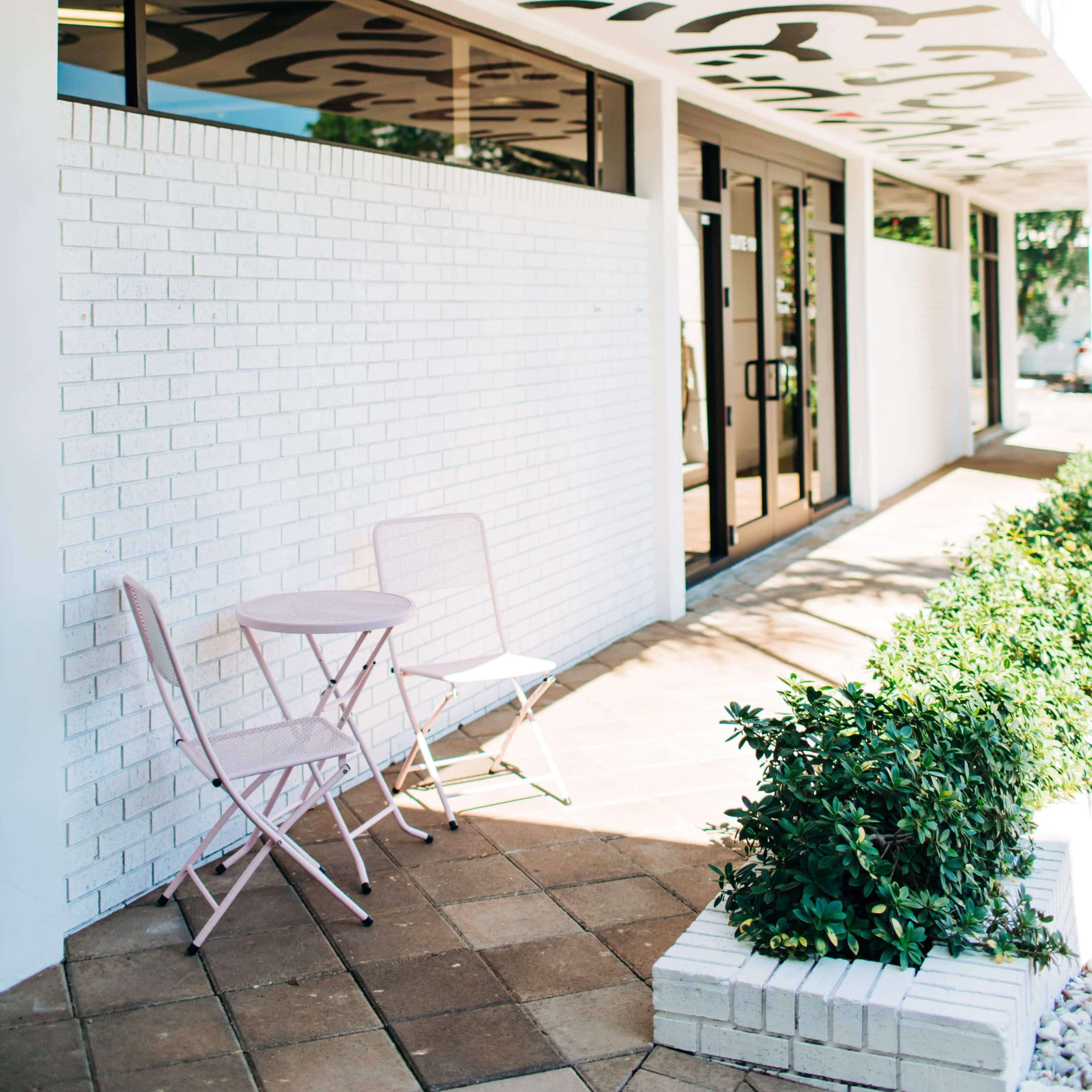 A small outdoor seating area features a pink bistro table and chairs in front of a building with a patterned ceiling.