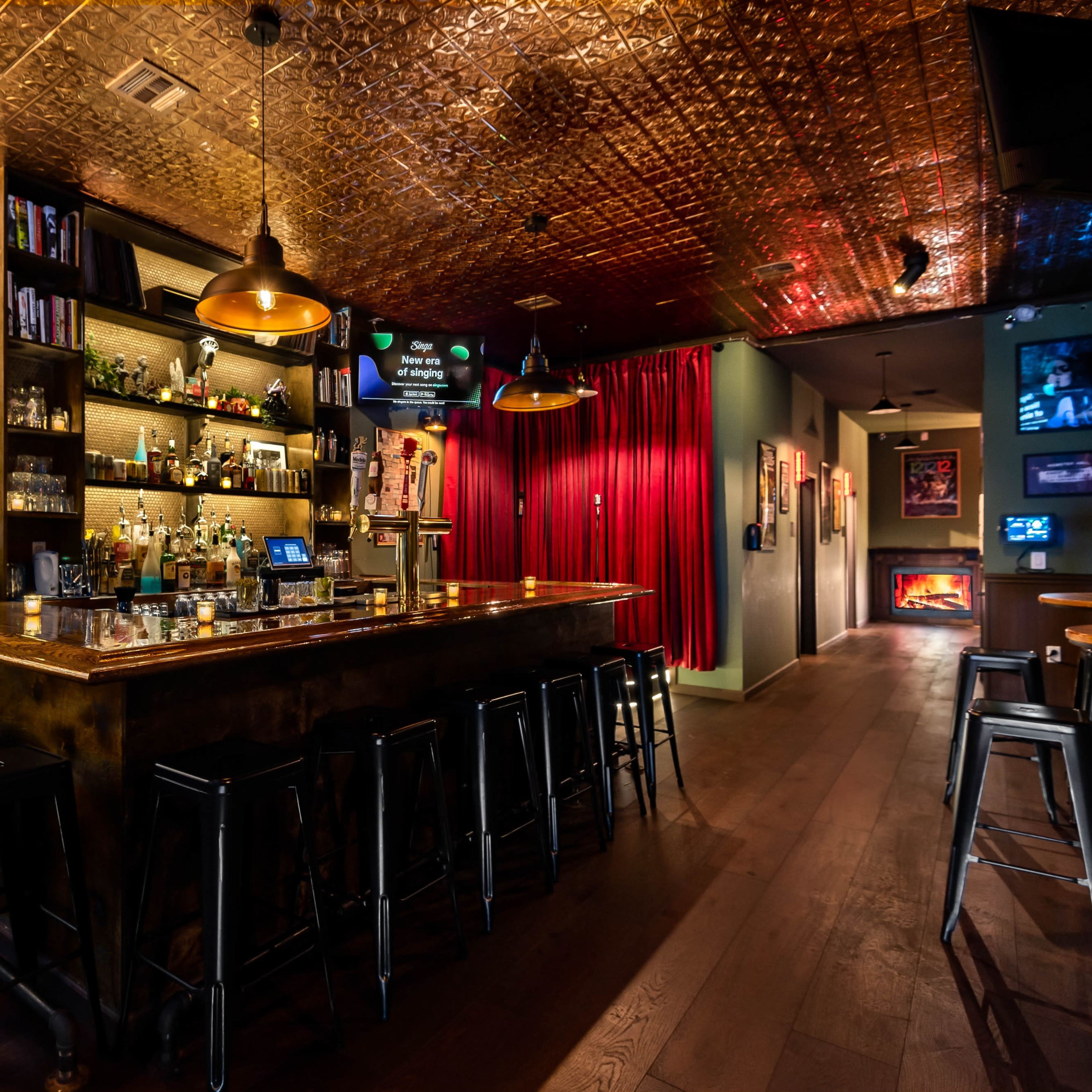 A dimly lit bar with a wooden counter, shelves of liquor, and several high stools arranged around it, leading to decorative curtains and illuminated art displays along a hallway.