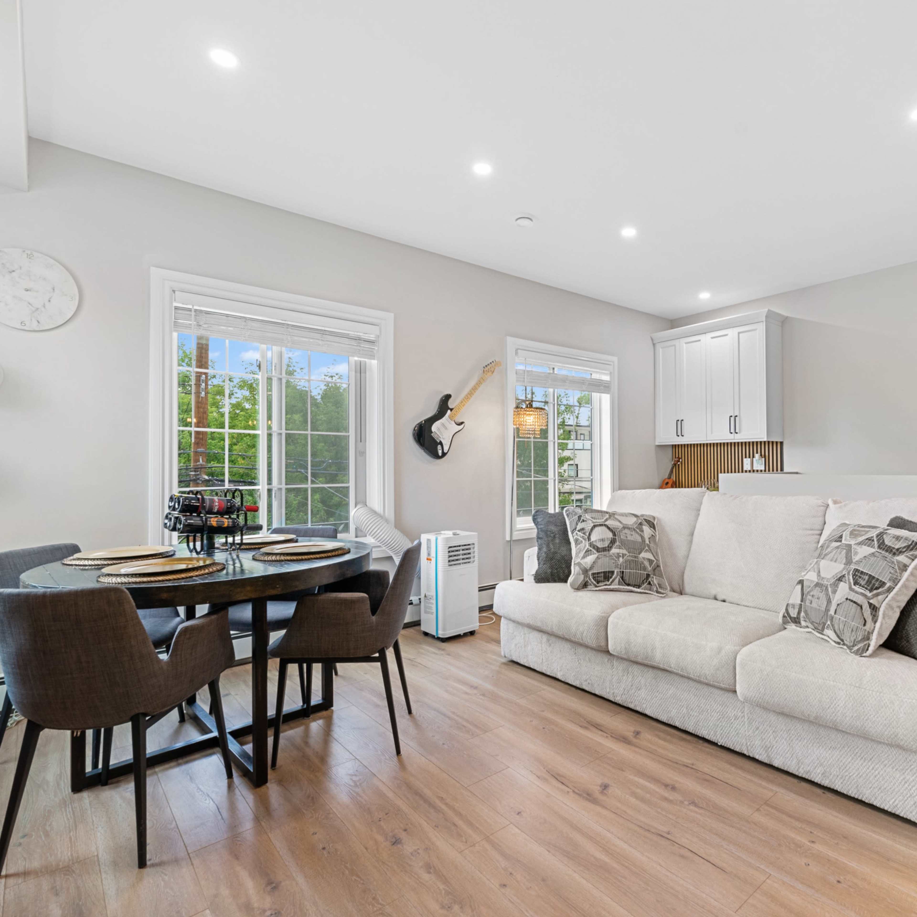 A modern living area featuring a white sofa, a round dining table with chairs, large windows, and a guitar mounted on the wall.