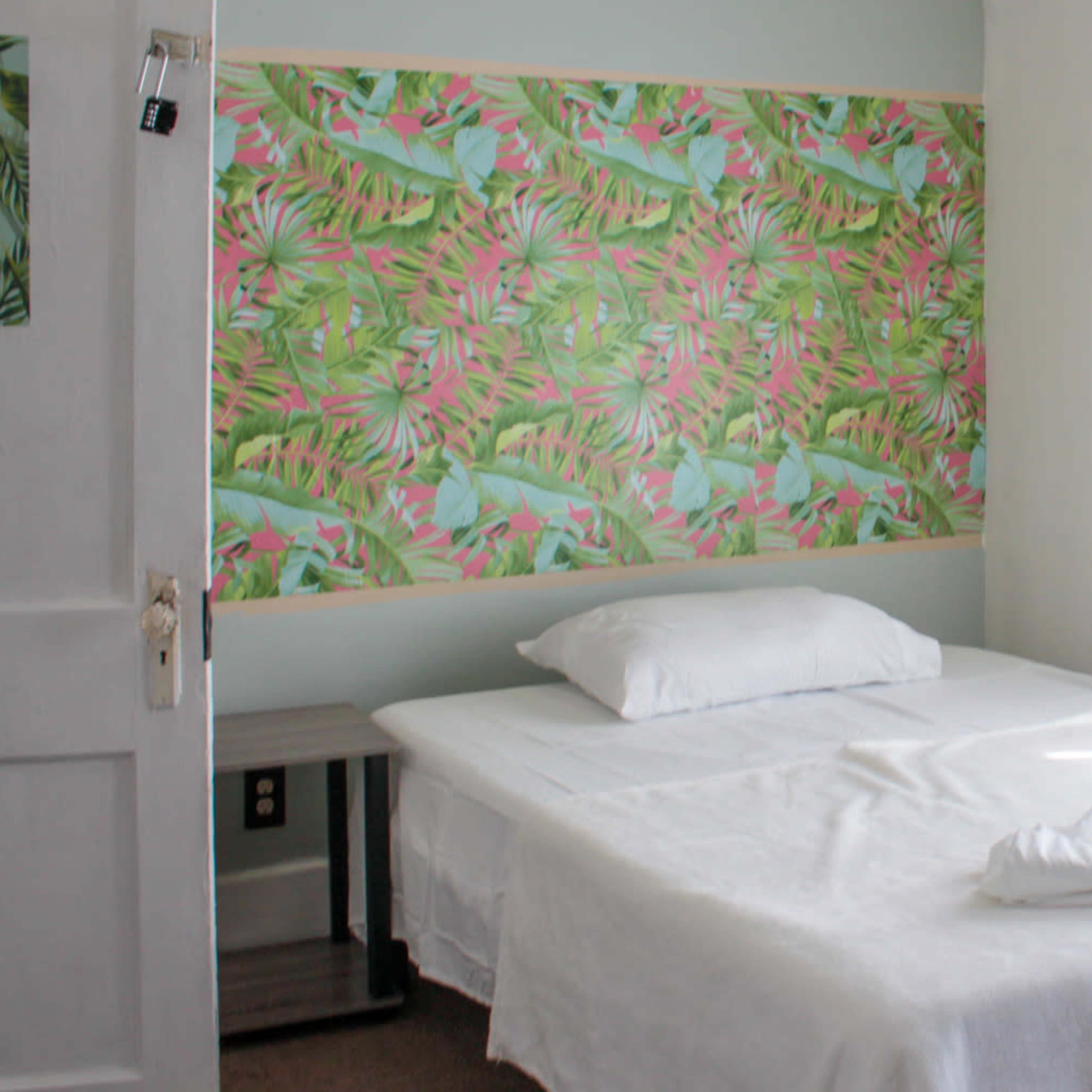 A simple bedroom with a white bed, a decorative wall featuring tropical patterns, and a window covered with sheer curtains.