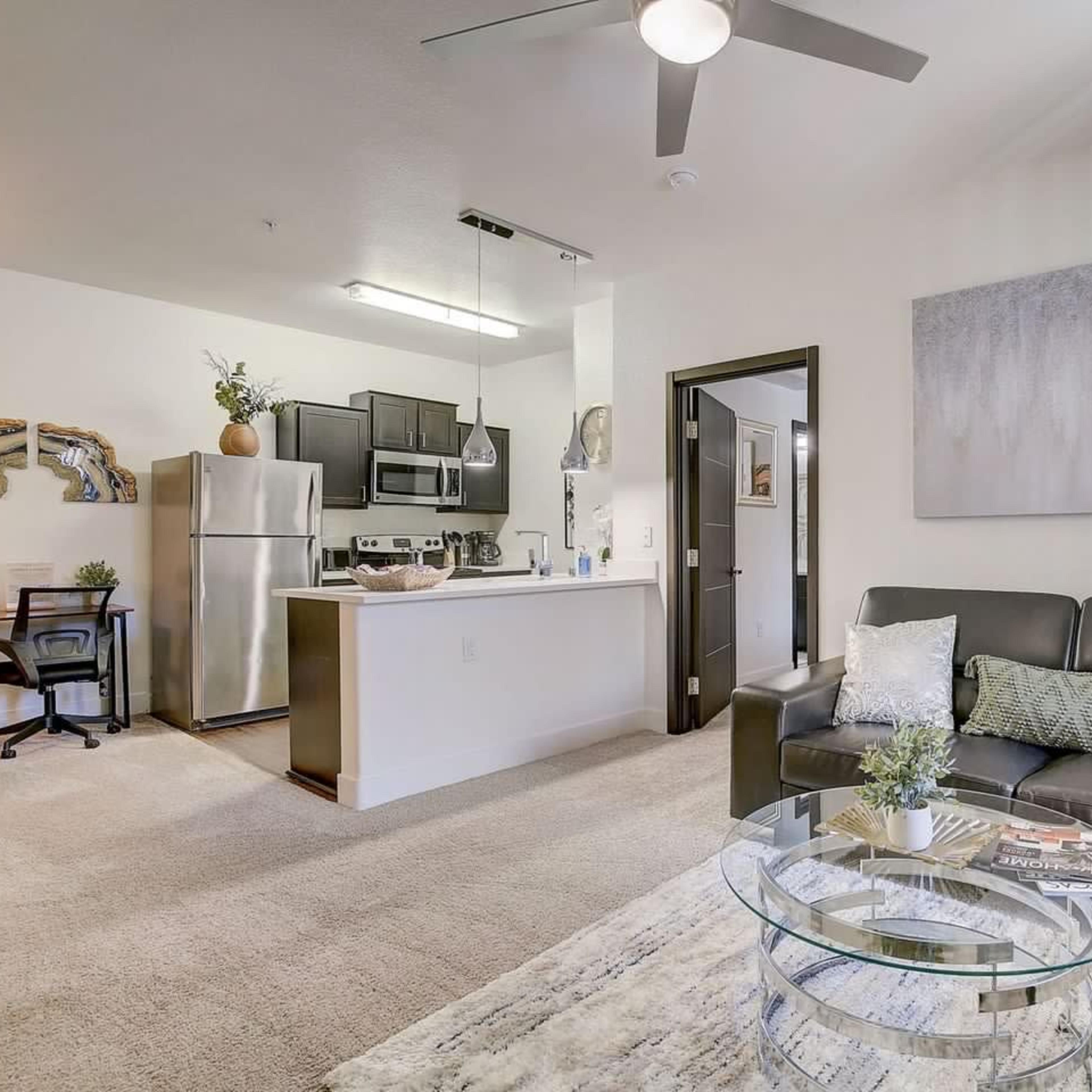 A modern living room with a black sofa, a glass coffee table, and a kitchenette in the background.