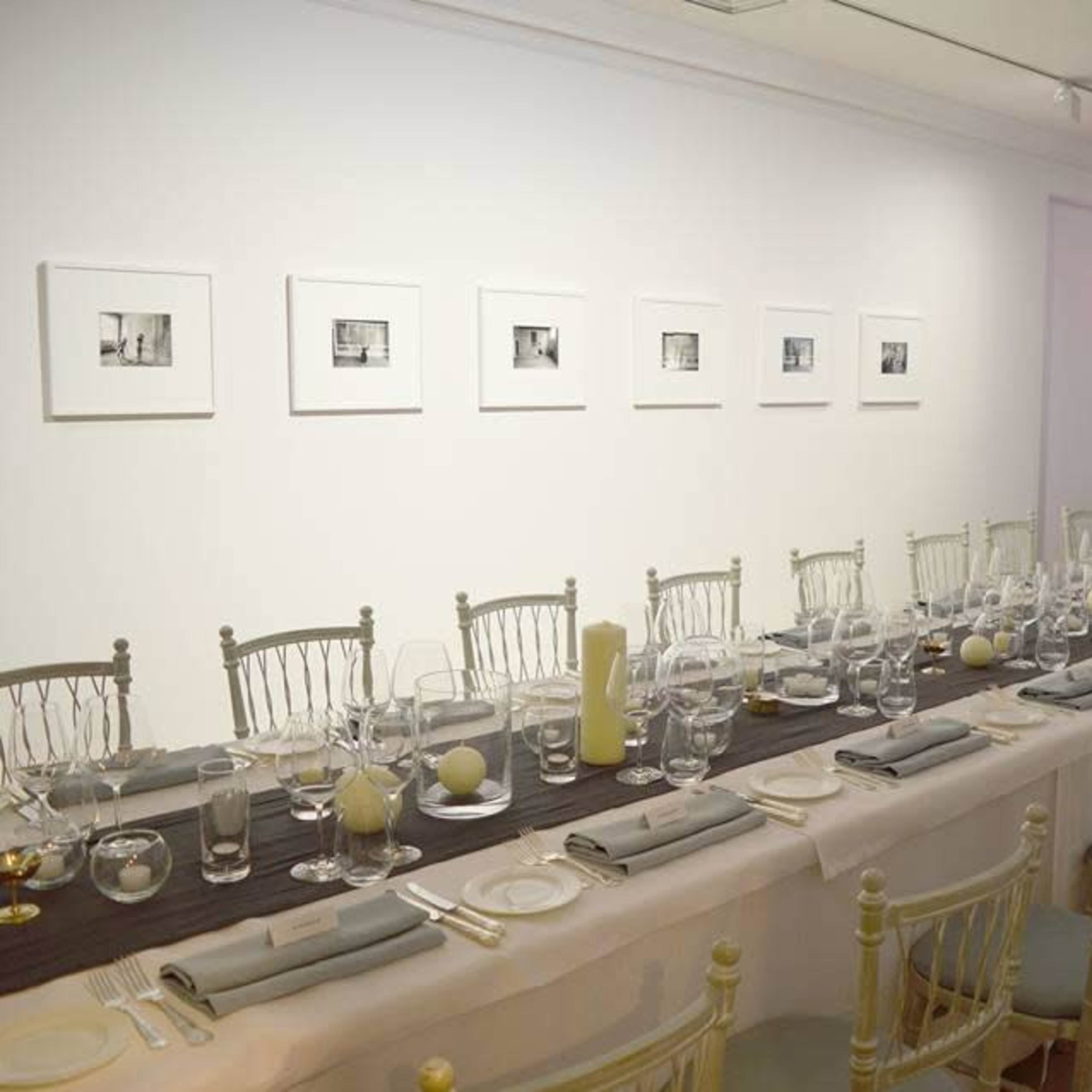A long, elegantly set dining table is placed in a bright room, flanked by framed black-and-white photographs on the wall and a large abstract artwork in the background.