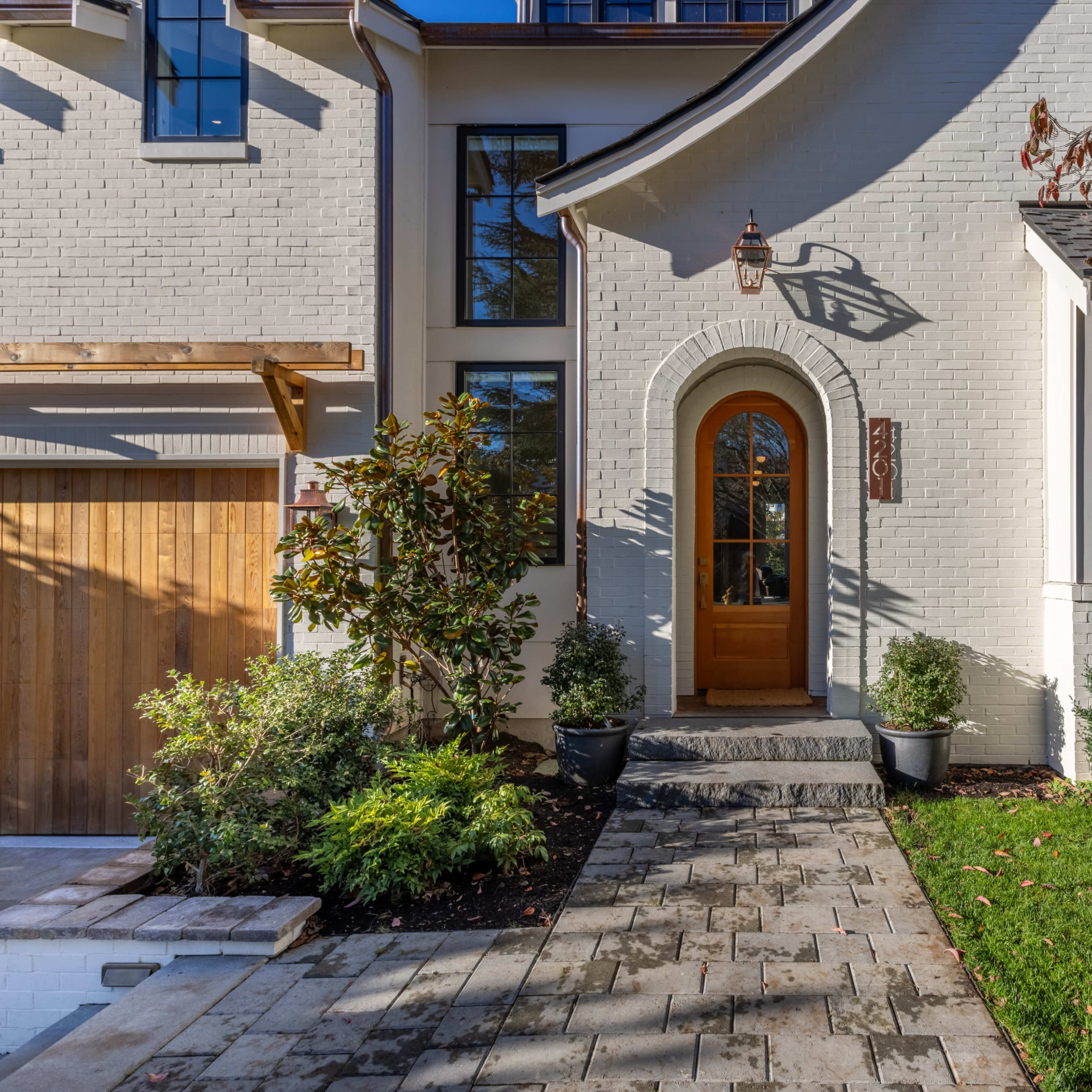 A well-maintained front entrance of a house with a wooden door, surrounded by greenery and a stone walkway leading to it.