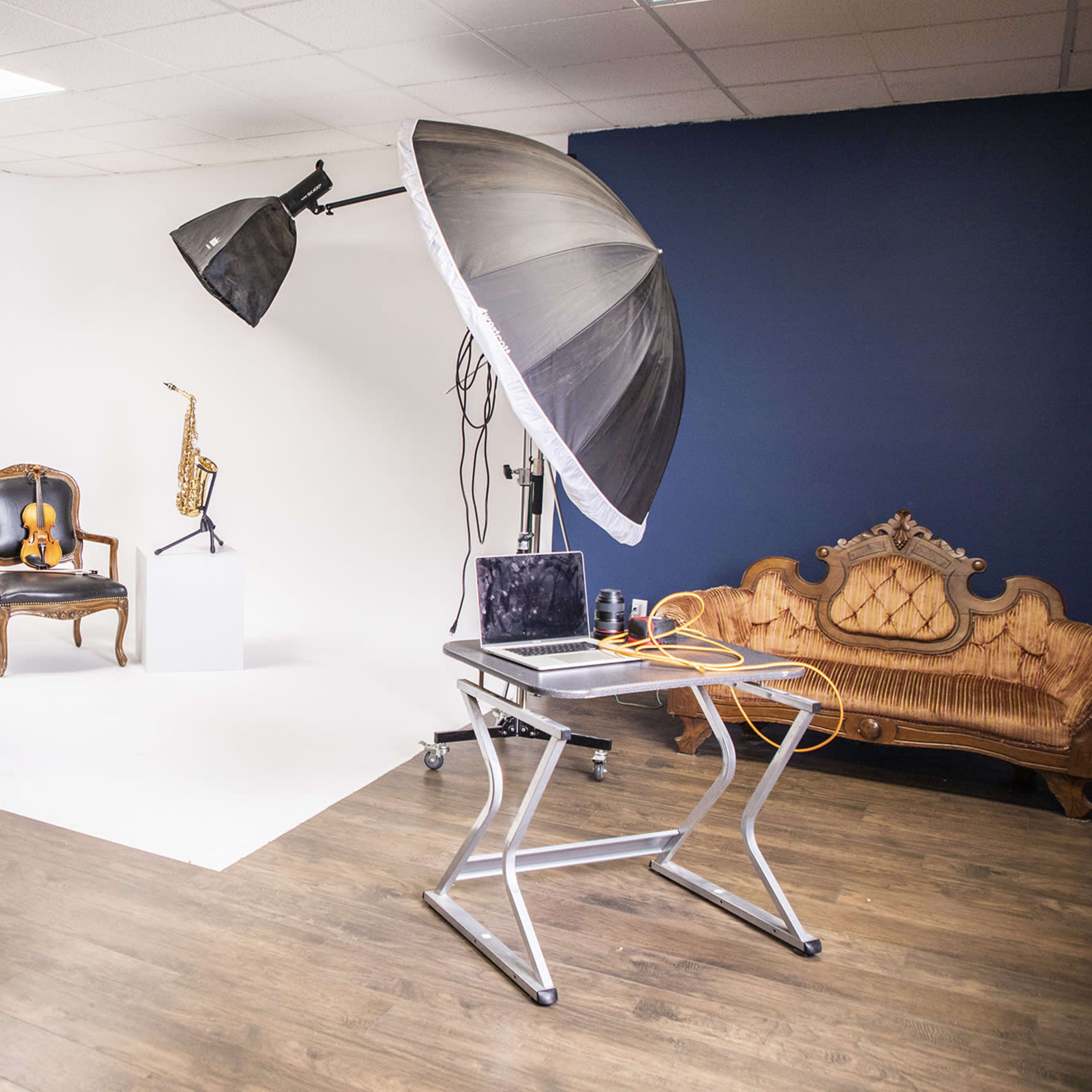 A photography studio setup with softbox lights, a camera on a tripod, a wooden chair, a saxophone displayed on a pedestal, and a laptop on a table.