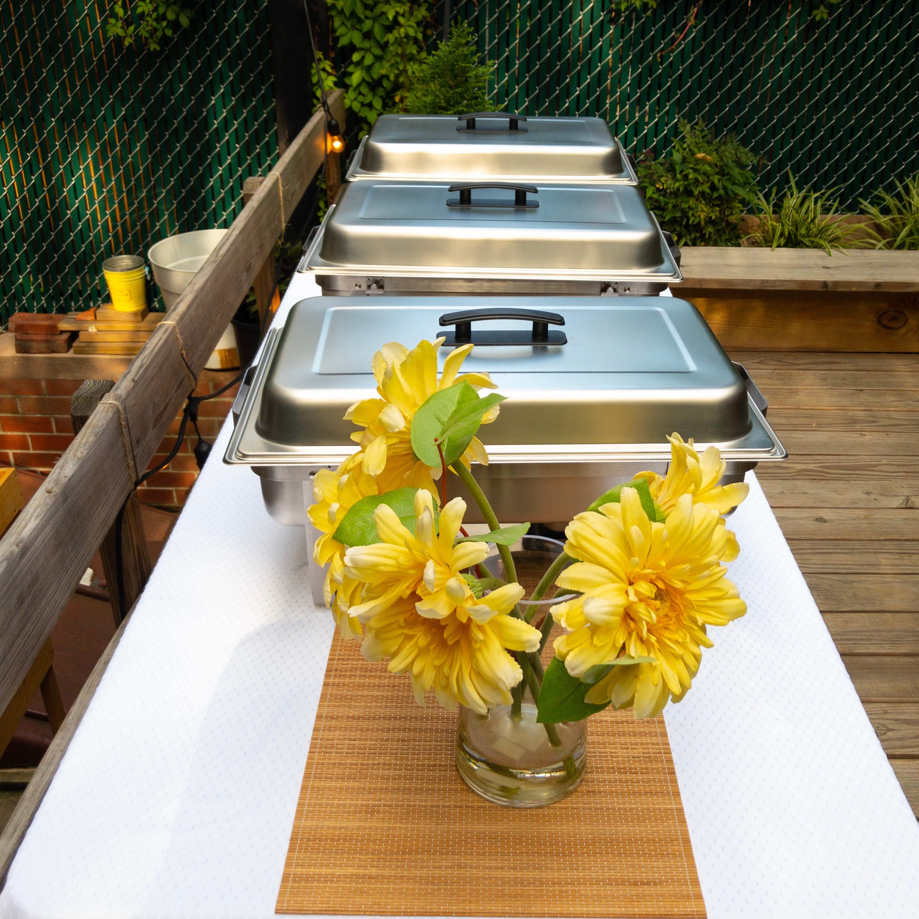 Three chafing dishes with lids are placed on a table alongside a vase filled with yellow flowers.
