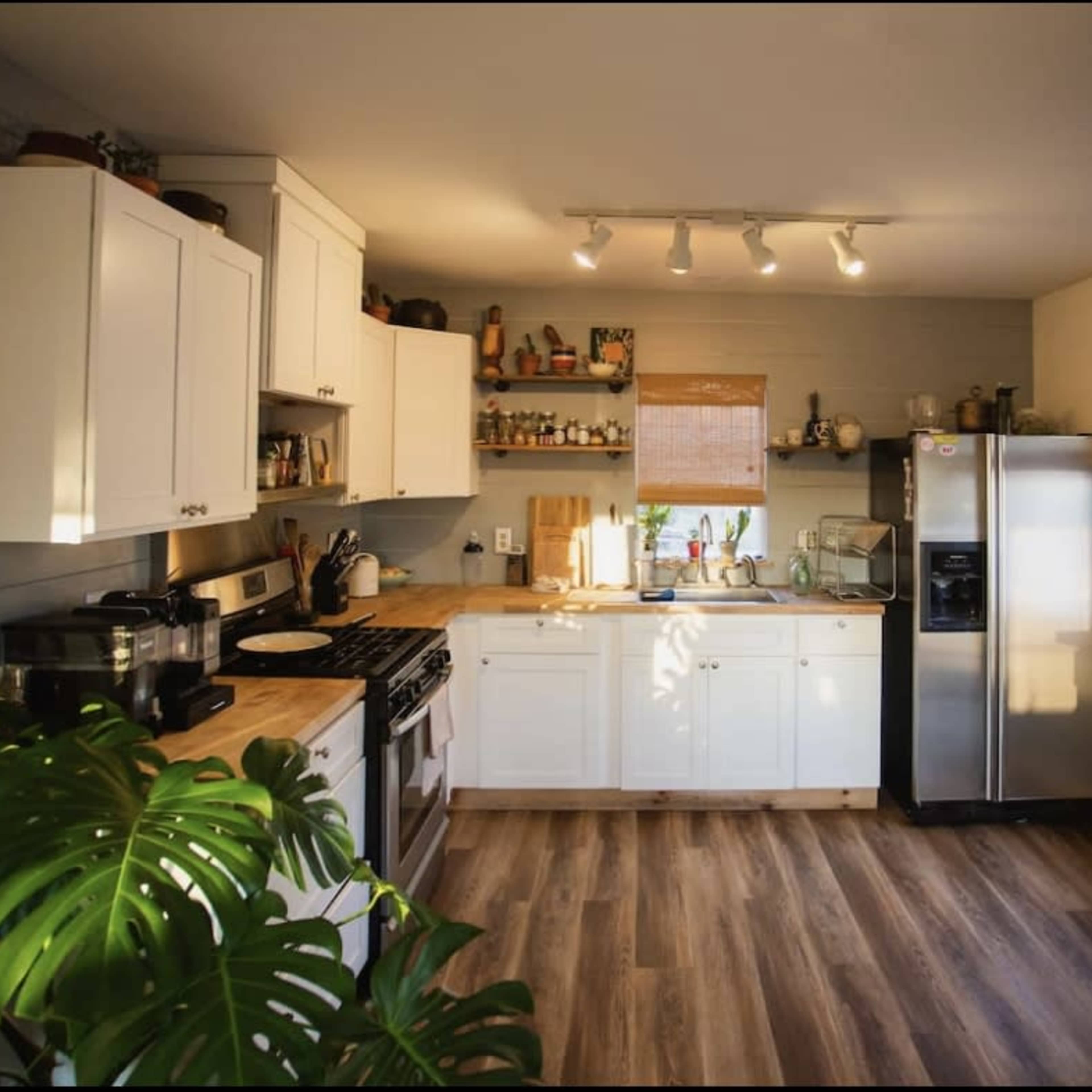 A well-lit kitchen features white cabinetry, a stainless steel refrigerator, and wooden countertops, with plants visible in the foreground.