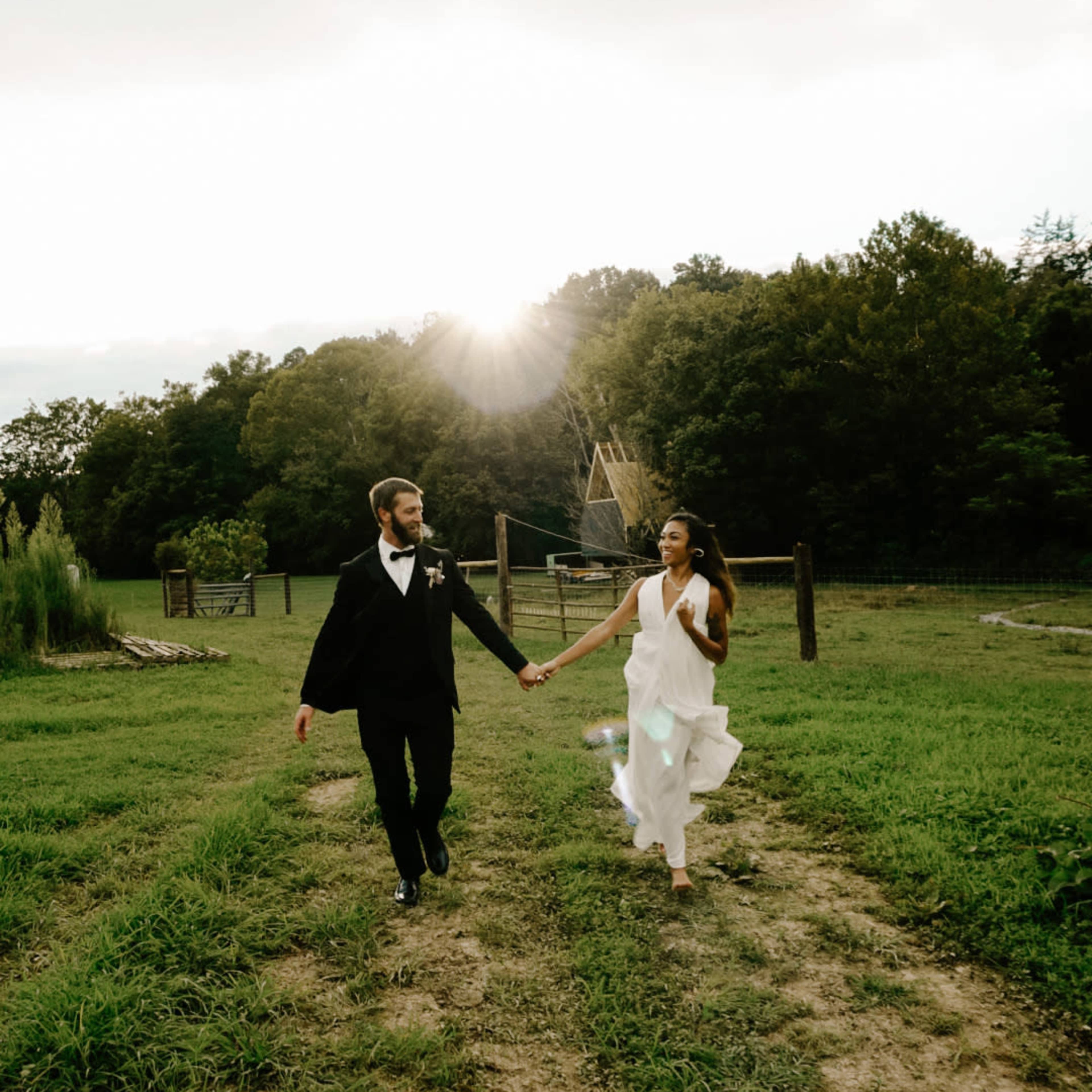 A couple in formal attire walks hand in hand down a dirt path in a rural setting with trees in the background and sunlight peeking through the clouds.