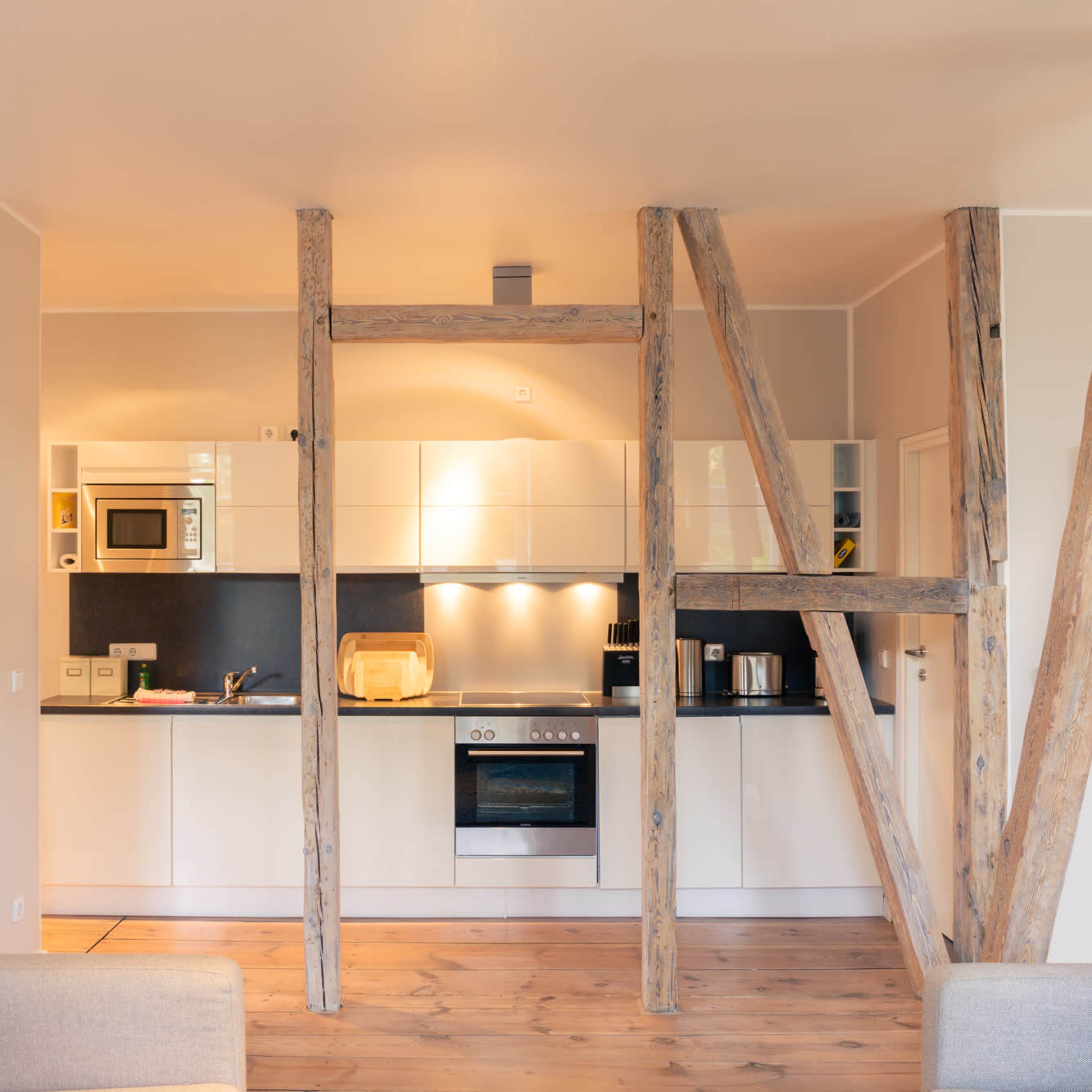 A modern kitchen with wooden support beams, featuring white cabinets, a stove, a microwave, and colorful artwork on the walls.