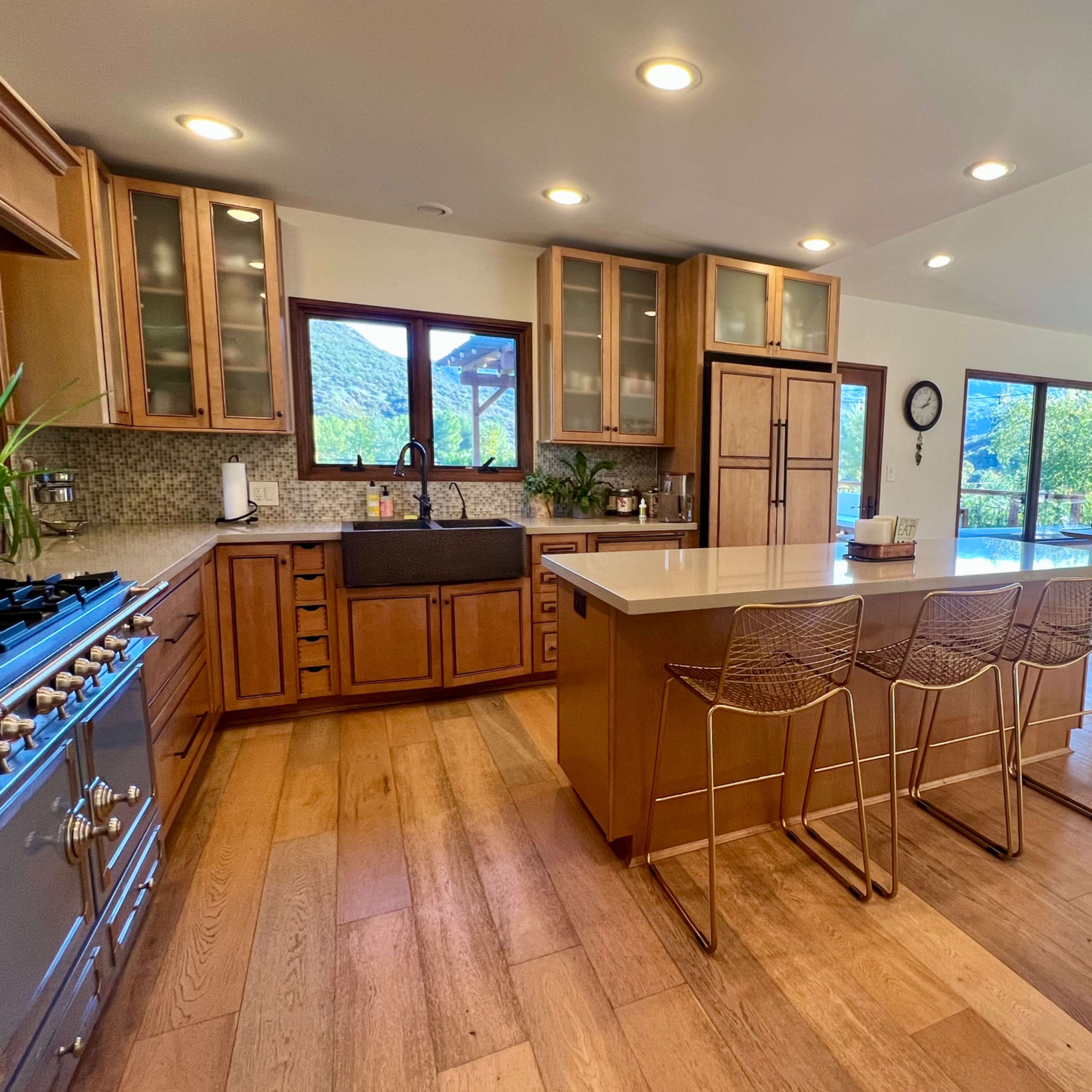 A modern kitchen features wooden cabinetry, a large island with barstools, and large windows overlooking a scenic view.