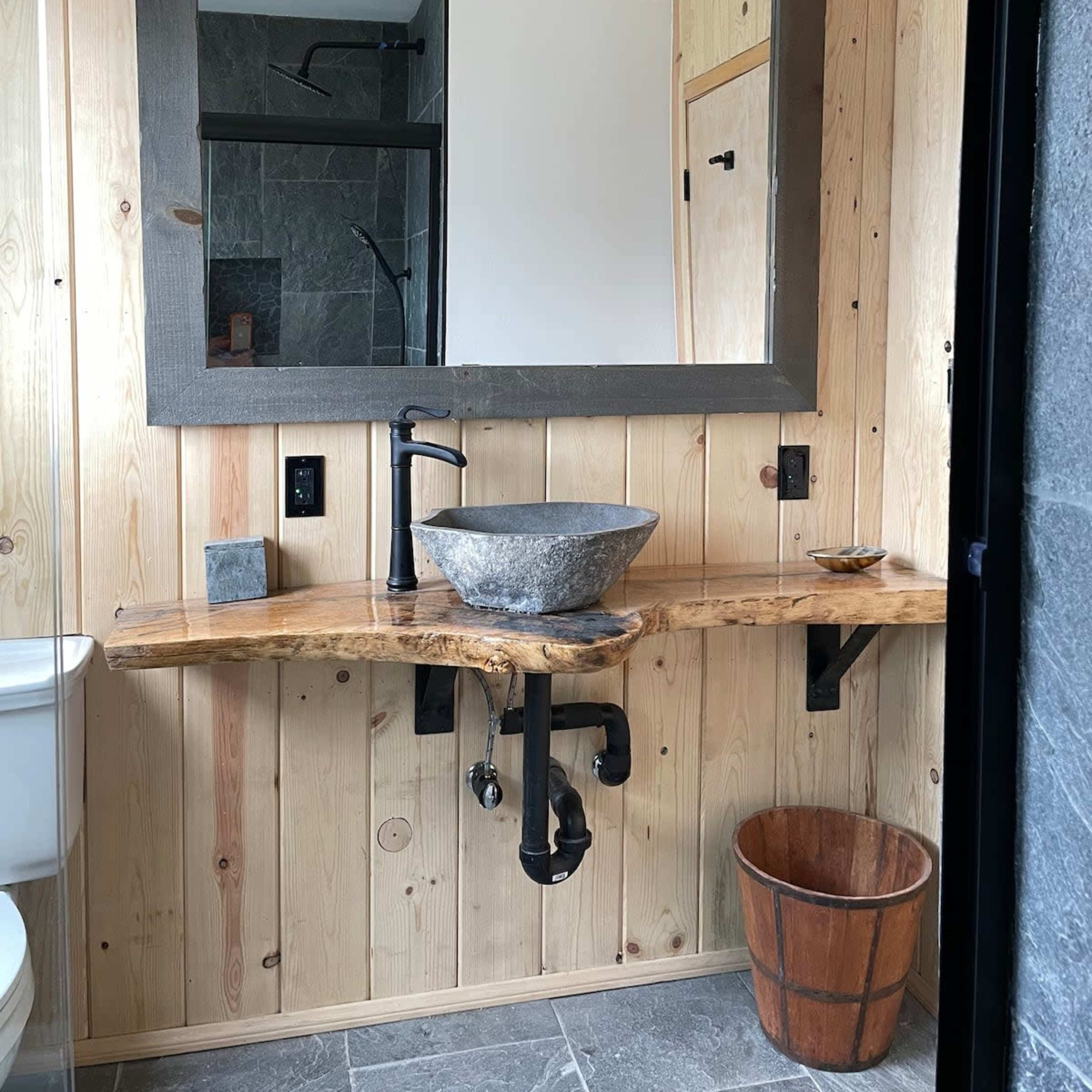 A modern bathroom with a wooden wall, a large mirror, a stone sink on a natural wood countertop, and industrial-style lighting fixtures.