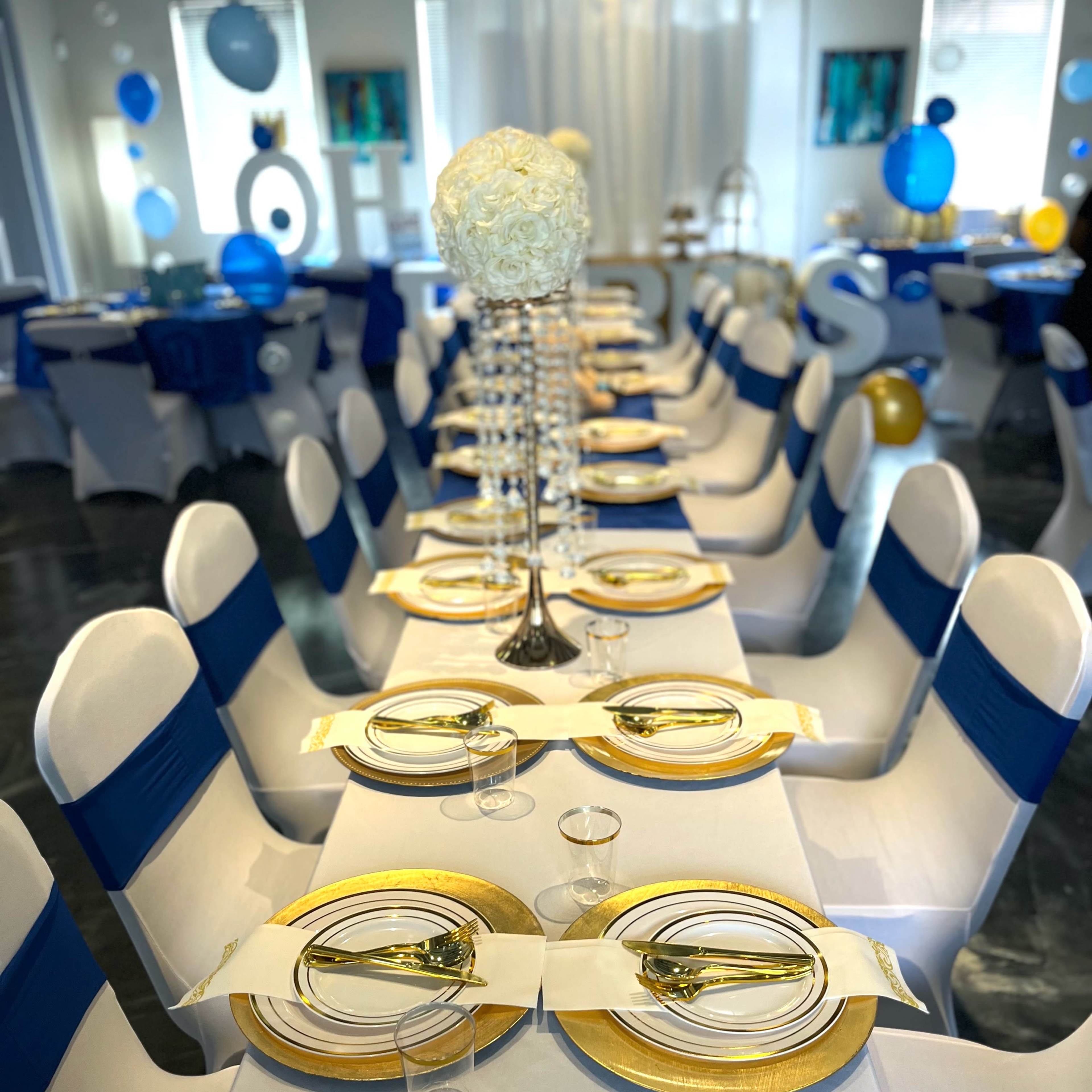 A banquet hall is set for an event with neatly arranged tables draped in white cloths and accented with blue bows, featuring gold rimmed plates and floral centerpieces.