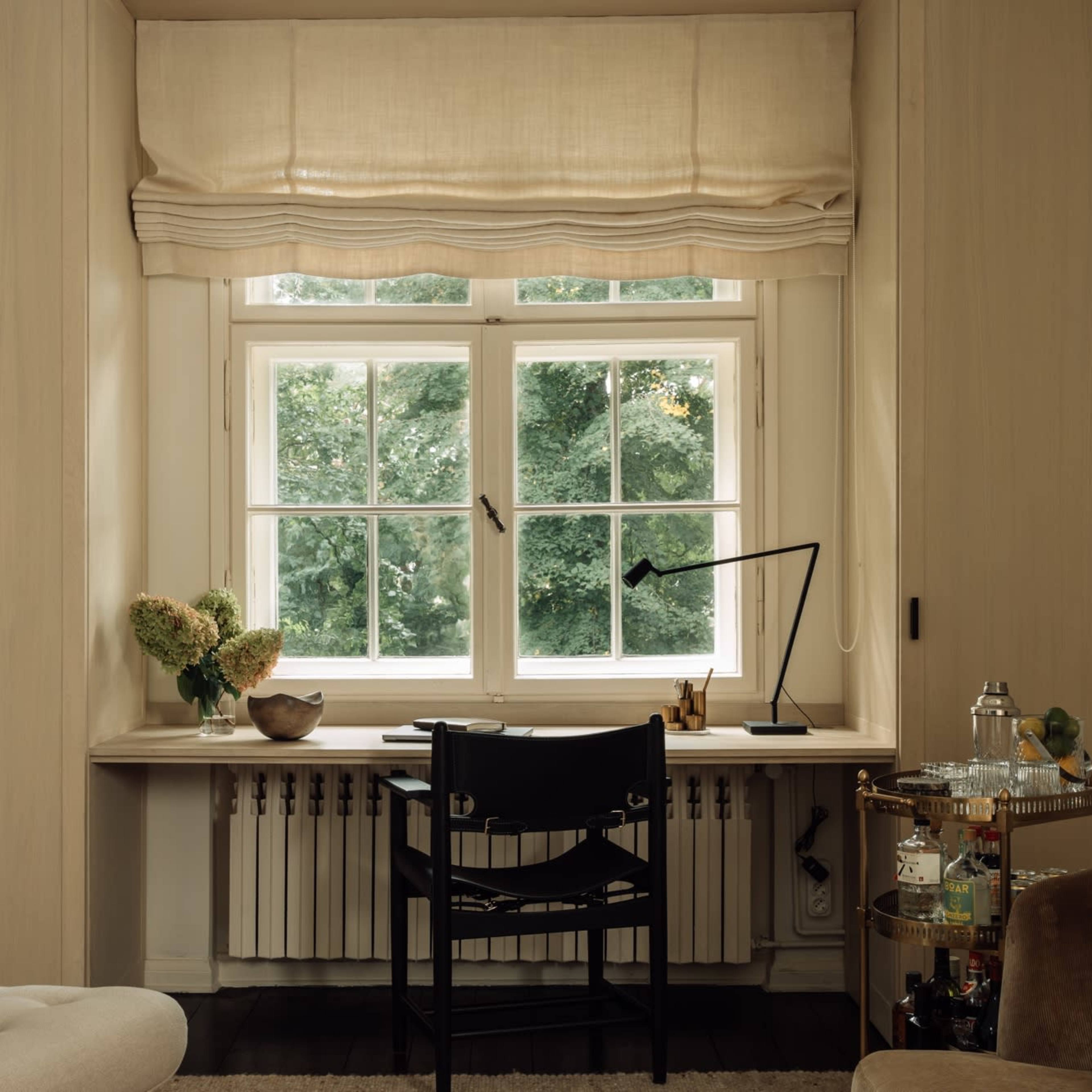 A minimalist home office features a desk with a black chair situated by a large window, overlooking greenery, with soft light filtering through a beige window shade.