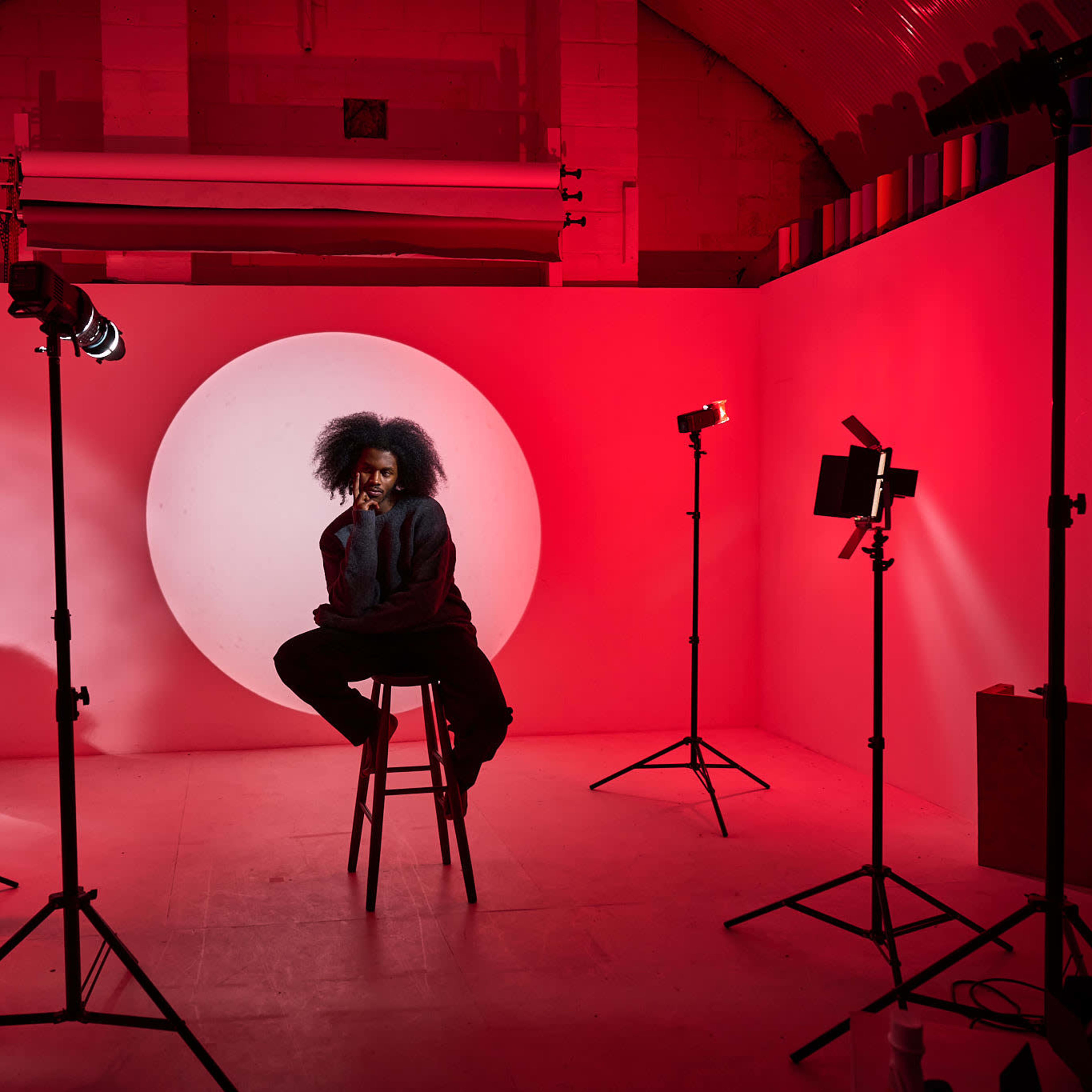 A person with styled hair sits on a stool in front of a large illuminated circular backdrop surrounded by studio lights.