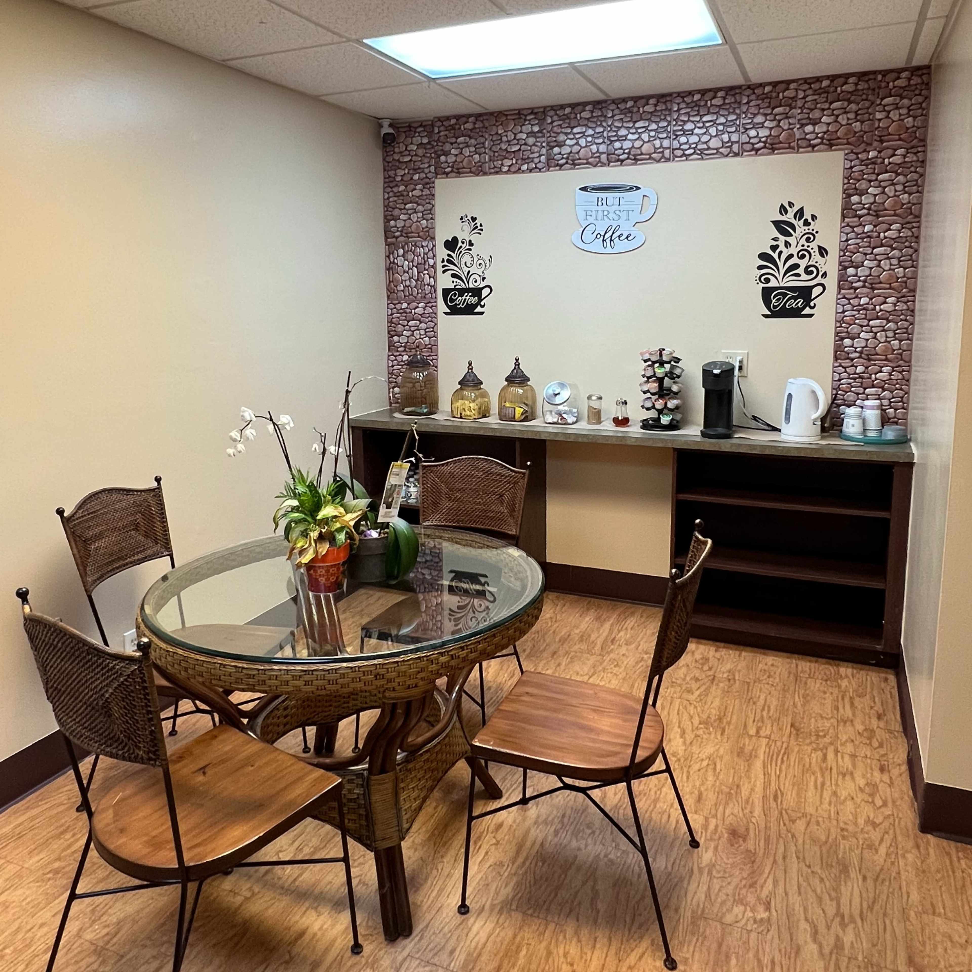 A small break room features a round glass table with four wooden chairs, a coffee station along the wall, and decorative wall art.