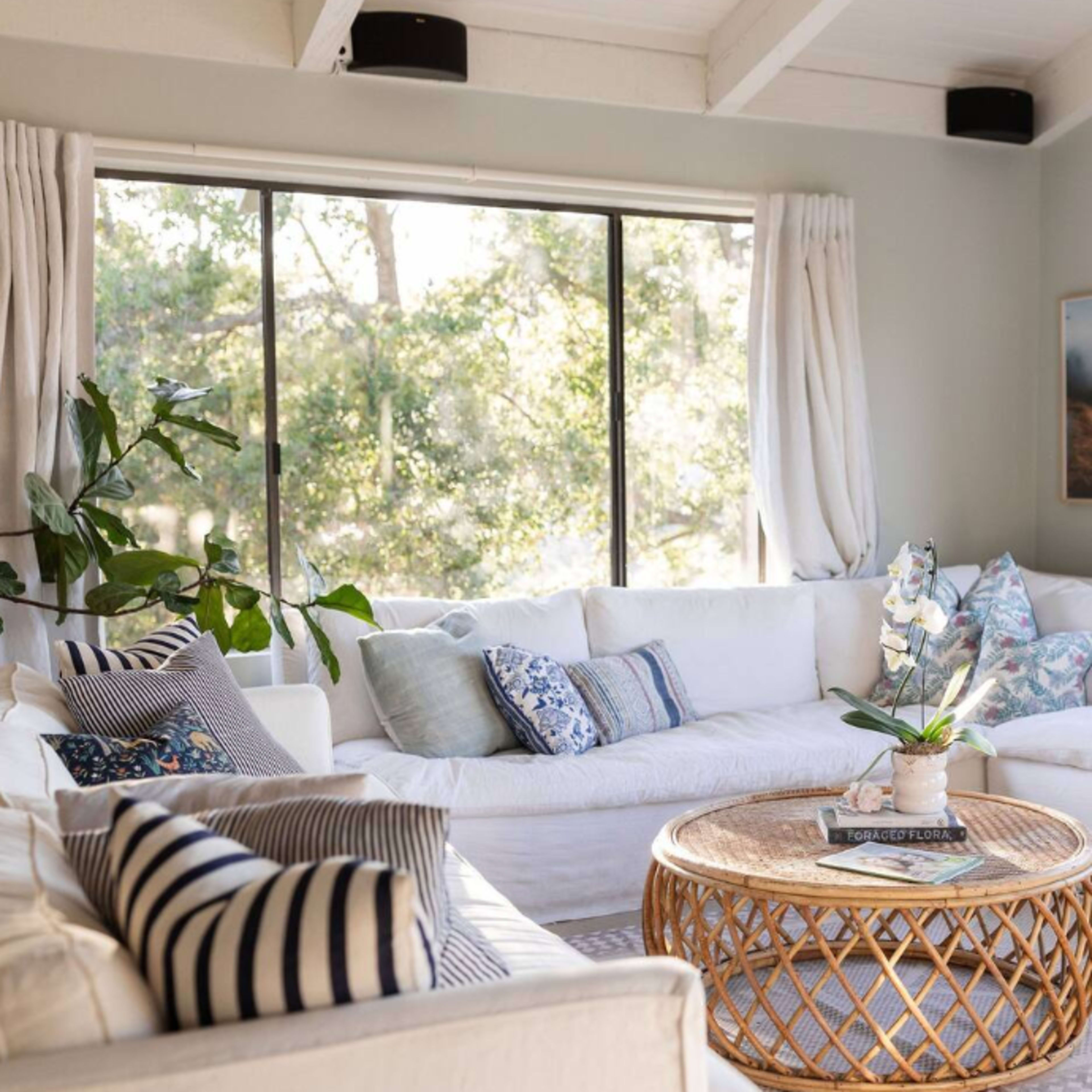 A bright living room with large windows, a white sectional sofa, a round rattan coffee table, and decorative pillows.