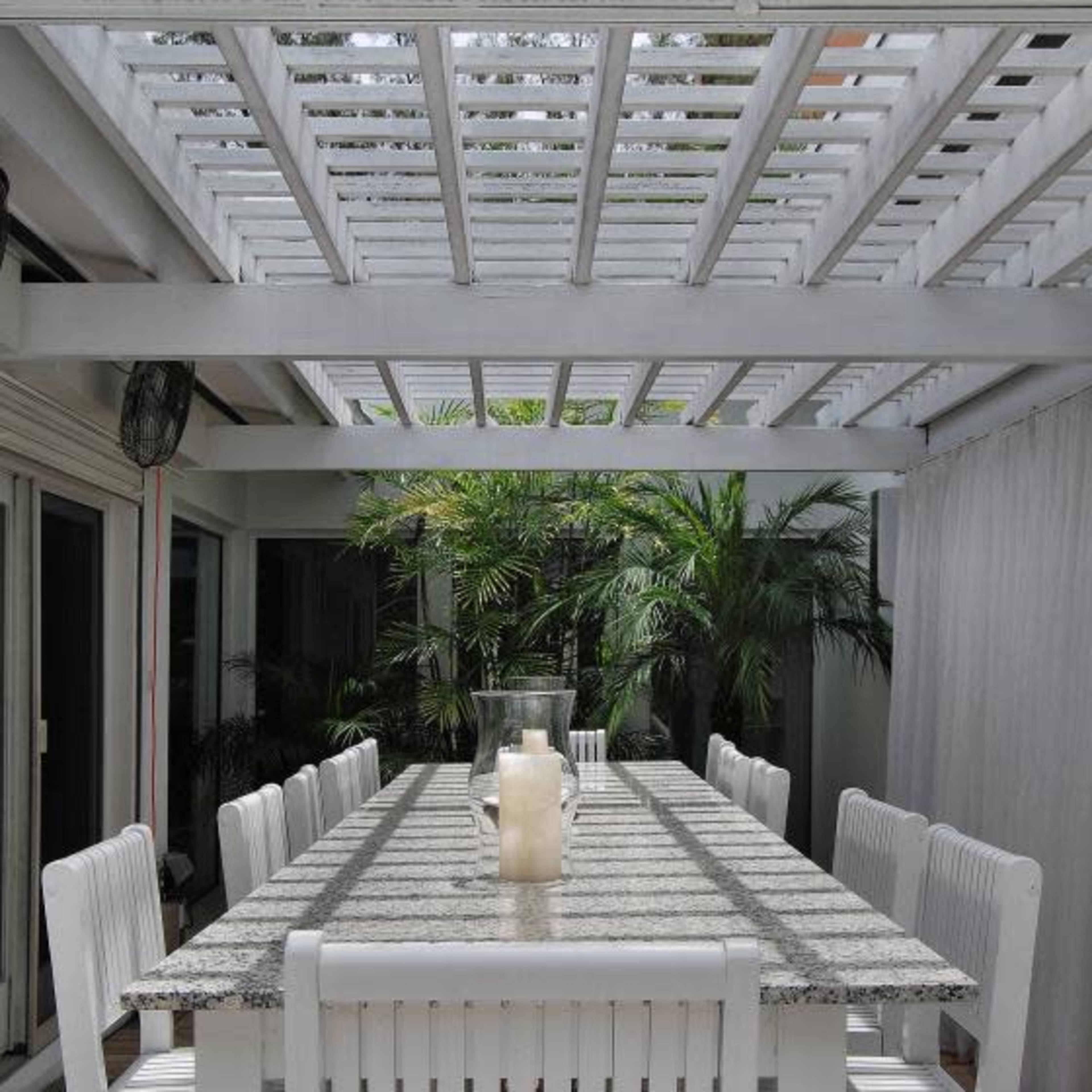 A long white outdoor table with high-backed chairs is set under a slatted wooden pergola, surrounded by lush greenery.