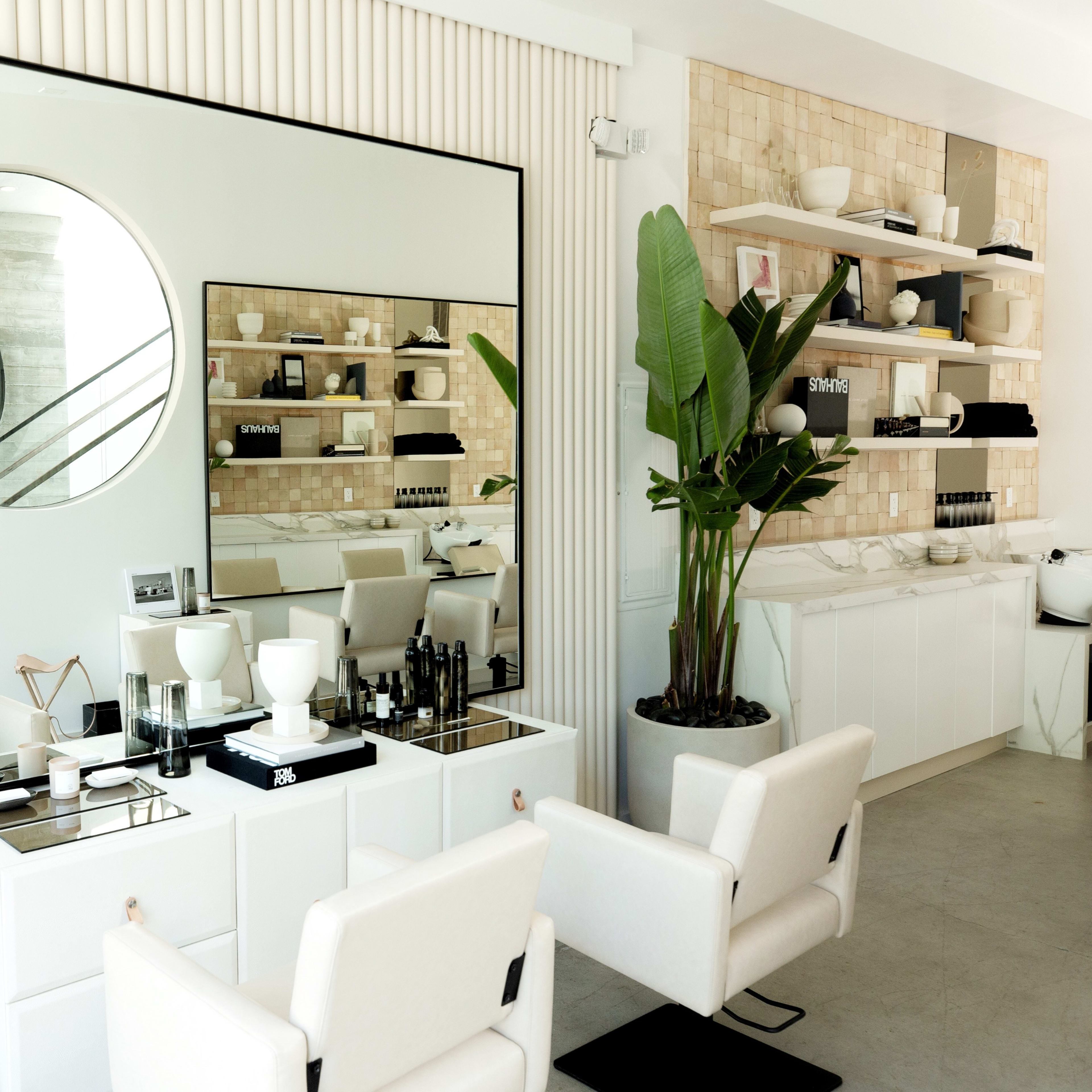 A modern salon interior featuring a large round mirror, two white salon chairs, and neatly arranged shelves with various beauty products.
