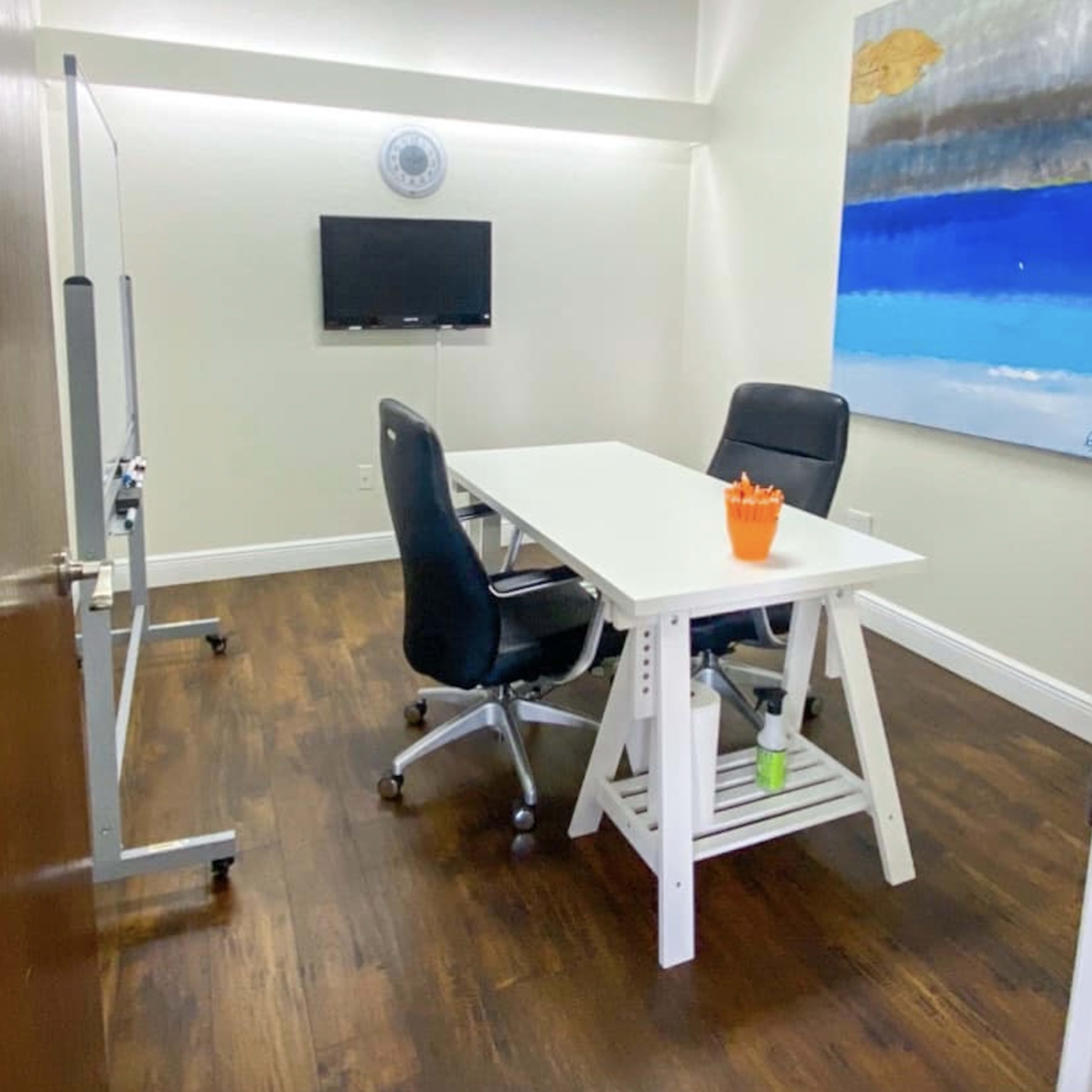 A small office featuring a white table, two black chairs, a wall-mounted TV, and a colorful abstract painting.